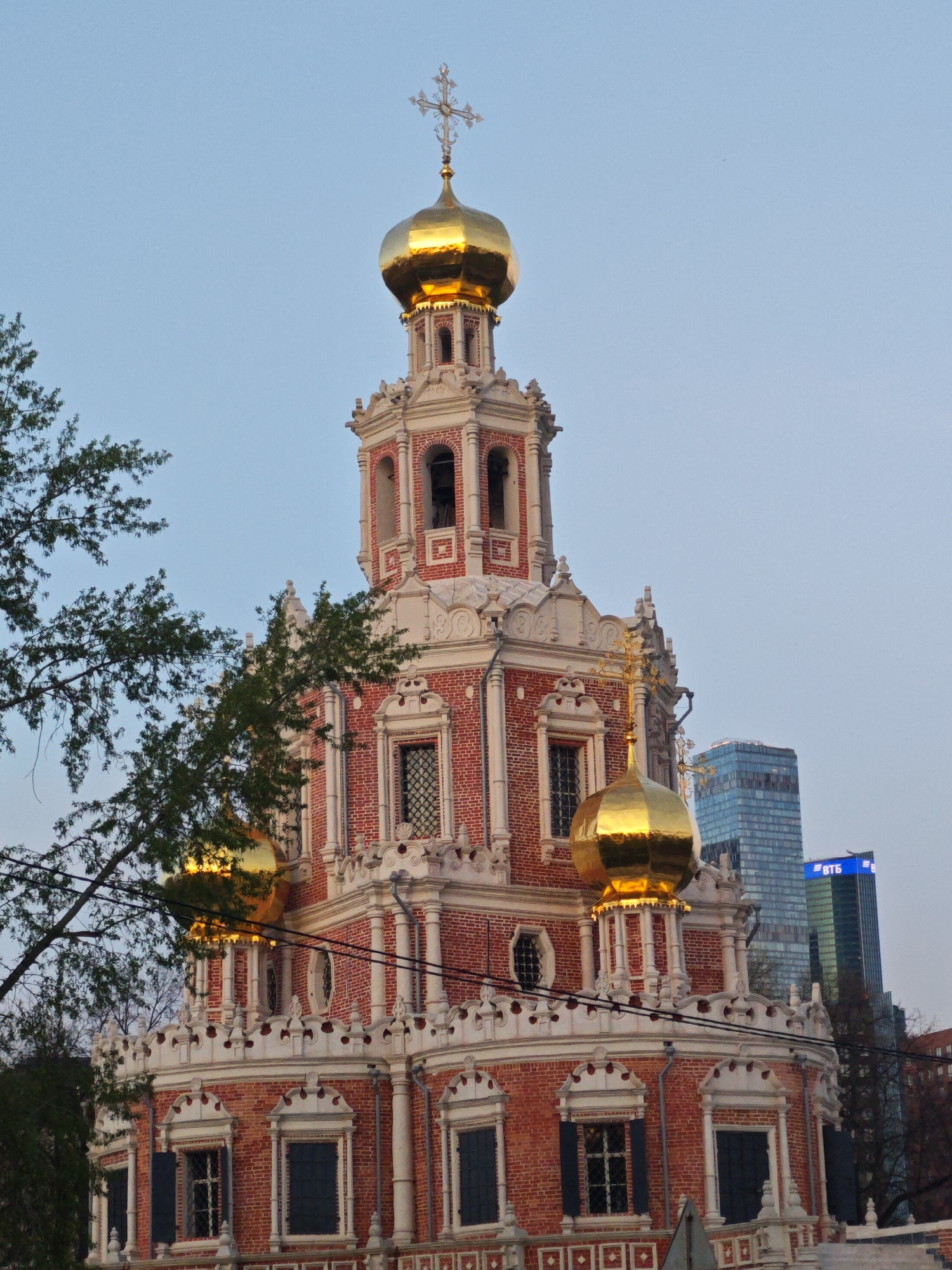 The image shows a building with golden domes and a cross on top. The building has a red and white brick facade with windows and decorative elements. It appears to be an Orthodox church. In the background, there is a modern skyscraper. The location is likely Moscow, Russia. The architecture of the church is distinctively Russian Orthodox. There are bare trees in the foreground, suggesting it might be autumn or winter.