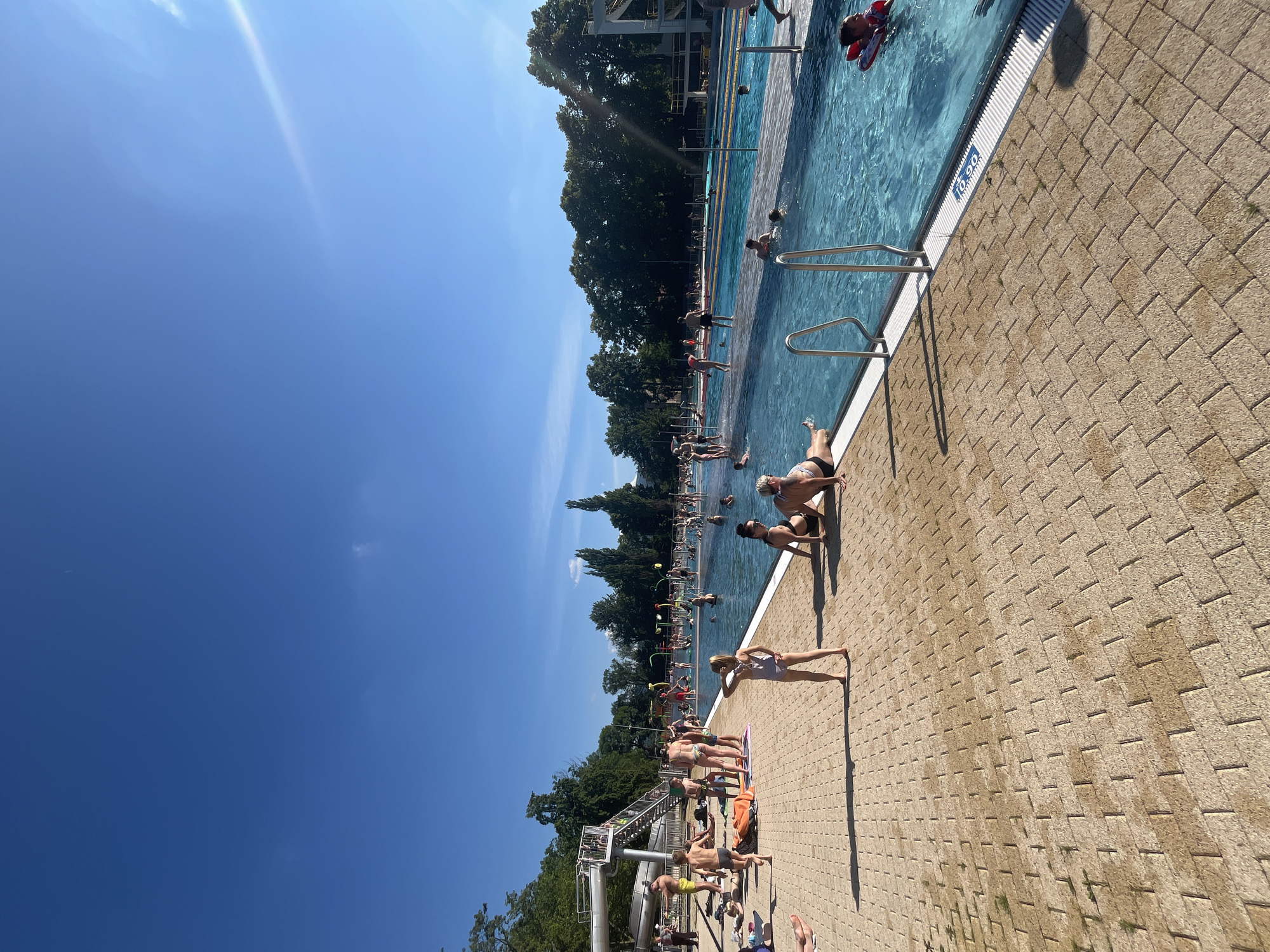 The image depicts a sunny day at an outdoor swimming pool. A clear blue sky dominates the upper portion of the frame, with a few wispy clouds visible. The swimming pool itself is filled with bright blue water, and several people are enjoying the water. Some are swimming, while others are lounging on the pool deck. The pool deck is paved with light-colored bricks and has several metal railings leading into the water.
In the foreground, several people are sunbathing on towels and the brick surface. One woman in a striped bikini is lying on her back, with her legs extended. Another person, wearing a purple swimsuit, is standing with their arms outstretched, appearing to be enjoying the sun. Further back, more people are scattered around the pool, some sitting, some standing, and some in the water.
In the background, lush green trees line the edge of the pool area, suggesting a park-like setting. A tall white structure, possibly a diving board or part of a larger complex, is visible above the trees. The presence of many people in swimwear and the bright, sunny weather indicate a recreational activity, likely on a warm day. The sun's angle suggests it is likely midday or early afternoon. A blue sign with white numbers "10.90" is visible near the edge of the pool.