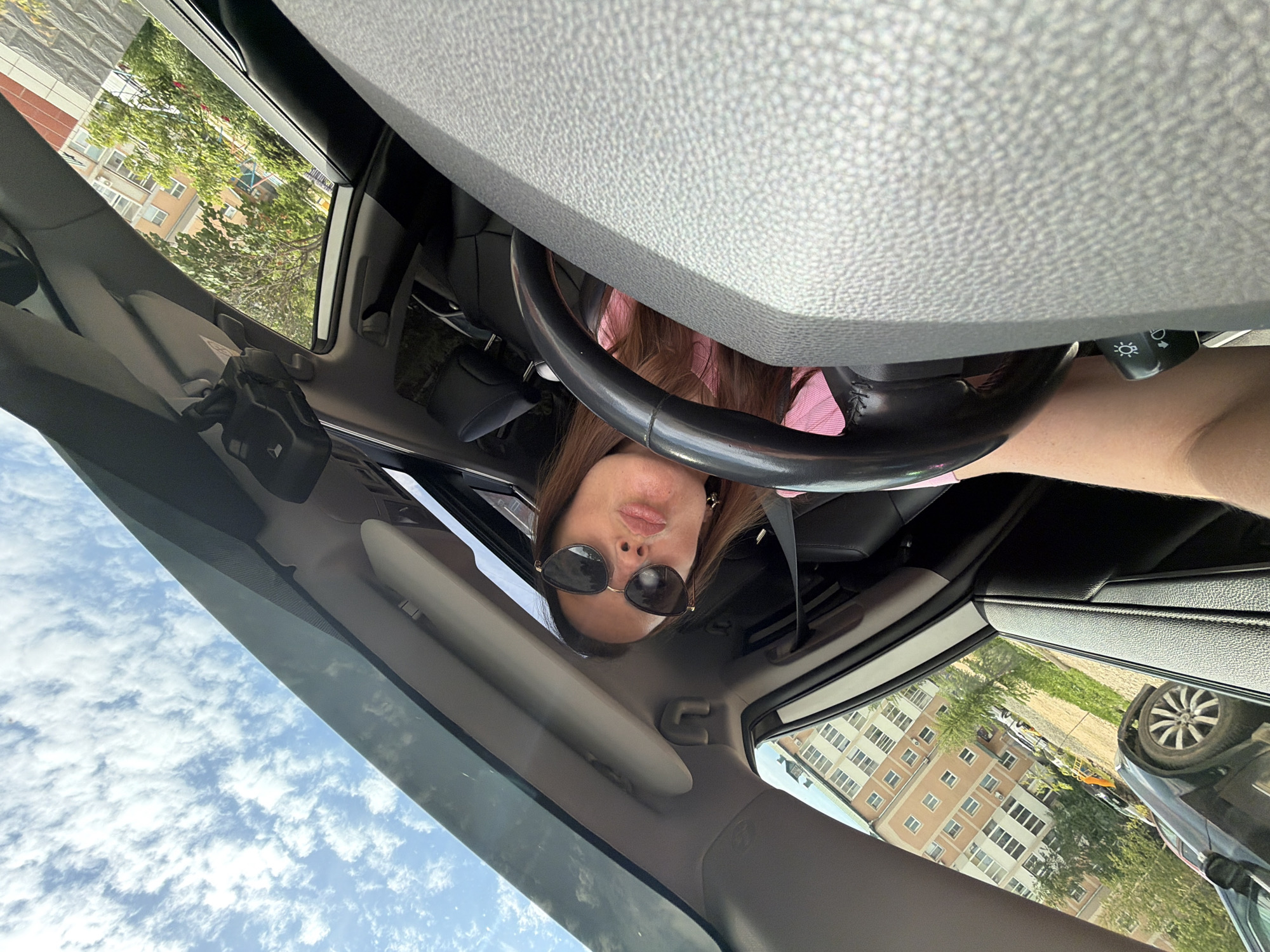 A first-person perspective selfie is captured from inside a car, looking towards the driver and the open sunroof. A woman, wearing sunglasses and a pink top, is seated in the driver's seat, her lips pursed in a kiss. Her hand rests on the steering wheel, and a turn signal lever is visible. The car's interior, including the textured dashboard and a portion of the passenger seat, is in the foreground. Outside the car, a black vehicle is parked on the left, and a multi-story apartment building with an orange facade, surrounded by greenery and trees, is visible. The sky above is partly cloudy. The location is the city of Birobidzhan, Russia.