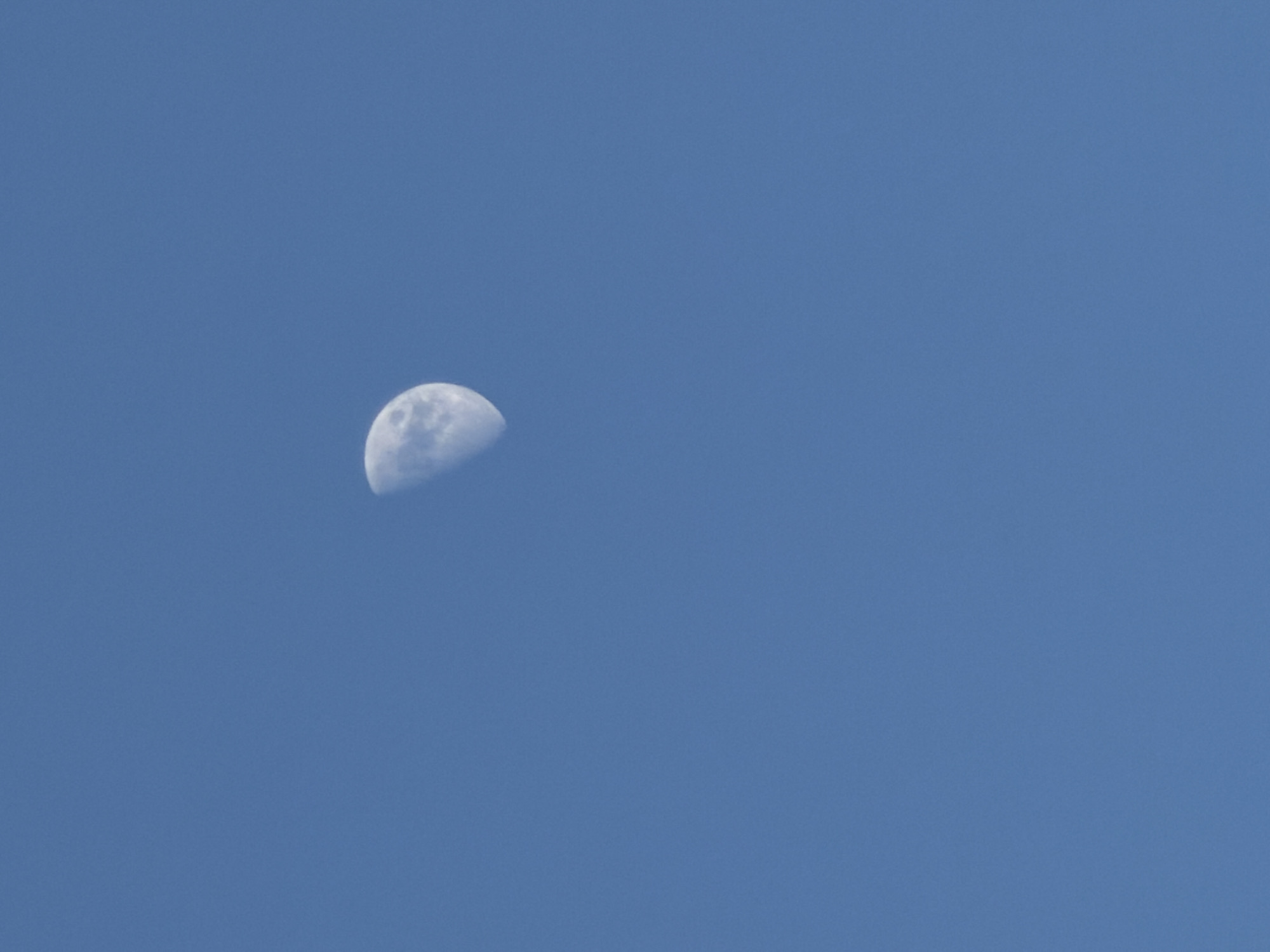 A photograph taken during the daytime shows a clear blue sky with a waning gibbous moon visible in the upper left quadrant. No people or cityscapes are present in the image. The location of the image is unknown, and cannot be determined as Moscow, Russia or any other city.