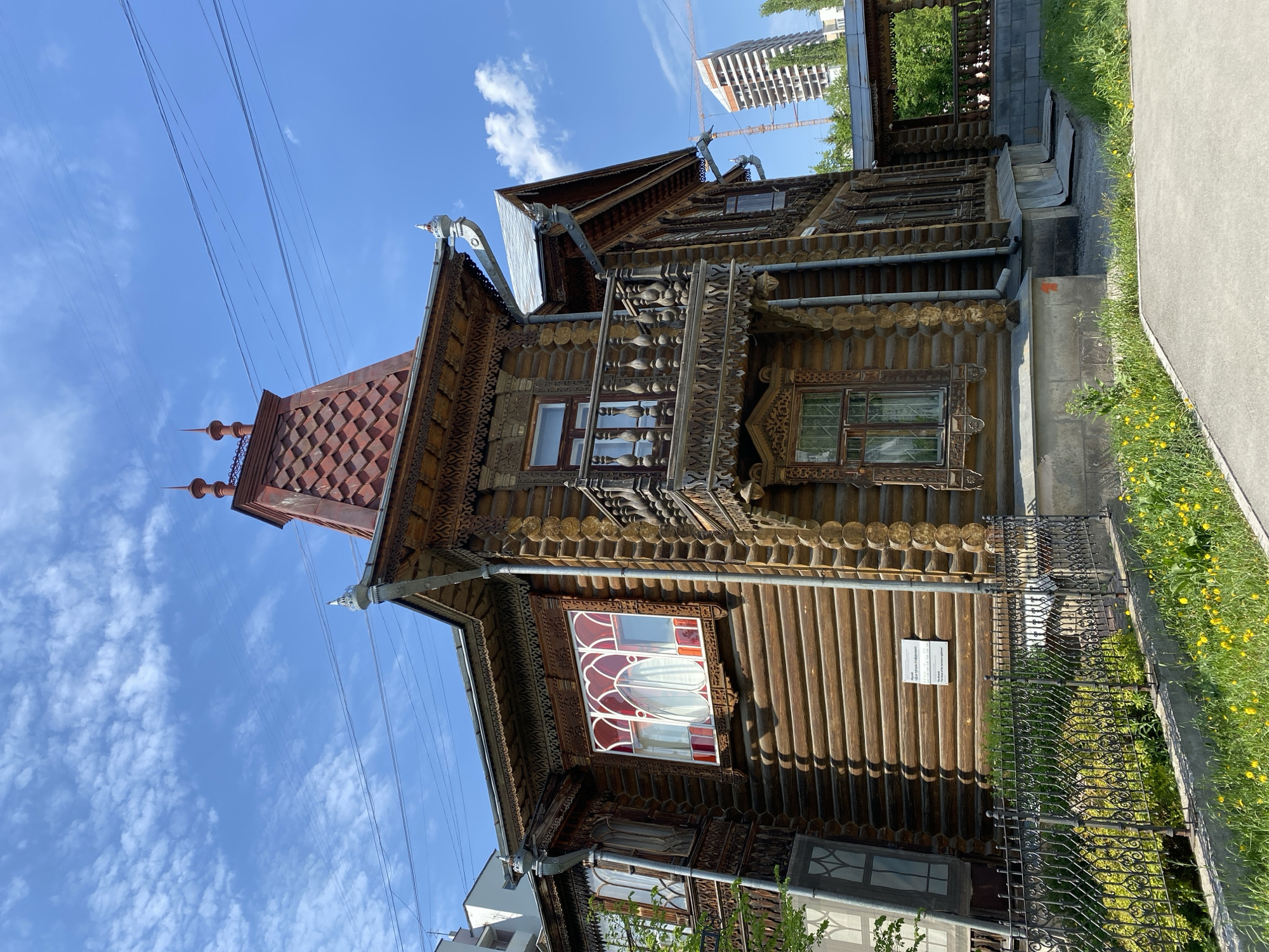 The image shows a detailed, ornate wooden house in Yekaterinburg, Russia, against a partly cloudy blue sky. The house is two stories tall, built from horizontal logs with intricate wood carvings adorning its windows, balconies, and roof. The roof has a distinct reddish-brown shingle pattern. A modern high-rise building is visible in the background behind the wooden structure. A black metal fence runs along the front of the house, separating it from a sidewalk with grass and yellow flowers along the edge. Power lines stretch across the upper part of the image. A sign is attached to the house's facade.