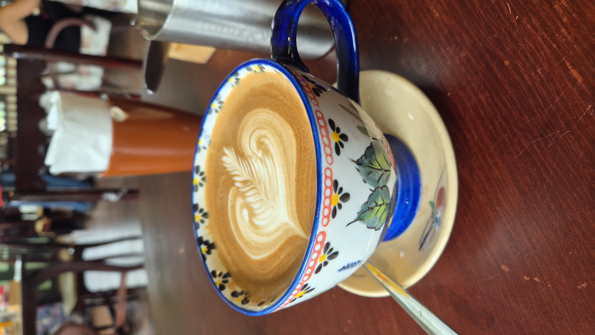 The image shows a close-up of a latte with intricate latte art, served in a decorative ceramic cup and saucer. The cup features a blue rim with yellow and black floral patterns, and green leaf designs on its sides, with a vibrant blue handle. A silver spoon rests on the saucer, positioned to the left of the cup. In the background, out of focus, is a blurred scene of a cafe or restaurant interior, with wooden tables and chairs, and other patrons present. A reddish-brown mug with white napkins is visible to the right of the latte. The overall setting suggests a casual dining environment, likely a cafe in Da Nang, Vietnam, given the context.