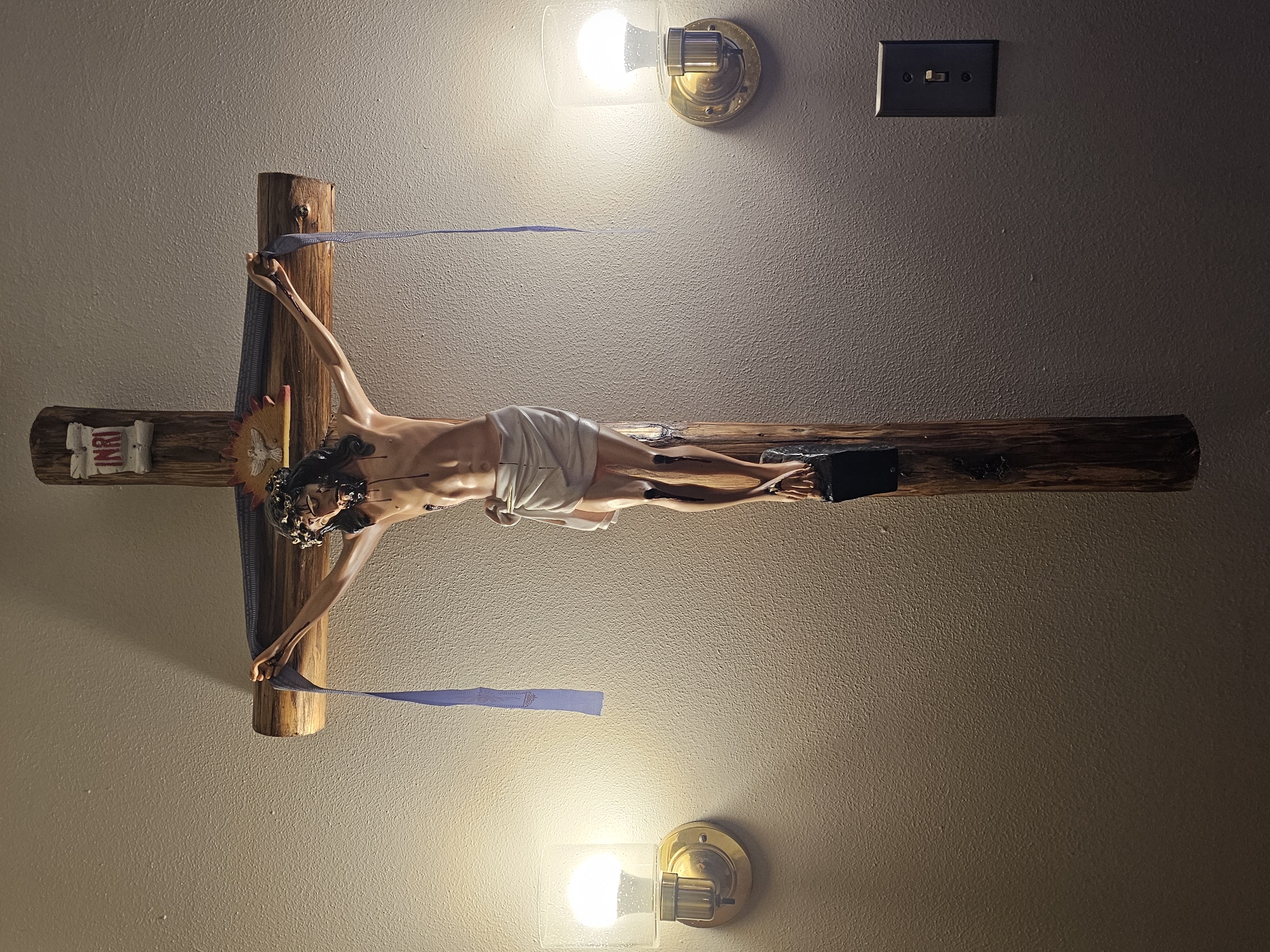 A crucifix hangs on a textured wall, illuminated by two wall-mounted sconces. The crucifix is made of rough wood and features a statue of Jesus in a loincloth, crowned with thorns. Two blue ribbons hang from the crossbars. The wall is a neutral color, with a light switch visible to the right of the crucifix. The scene is indoors, suggesting a religious or devotional setting. The location is identified as Amarillo, United States of America.