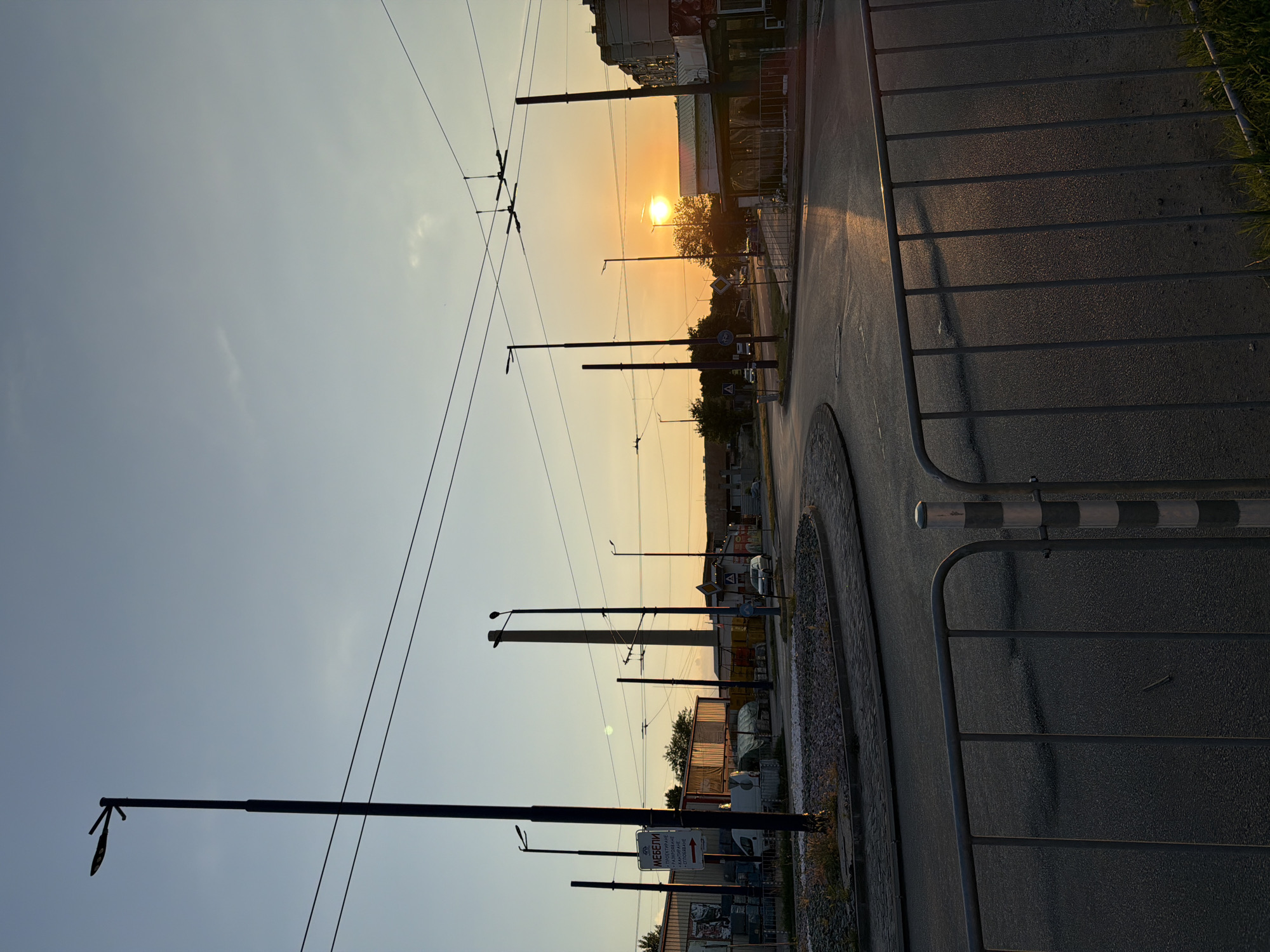 The image depicts a street intersection in Pazardzhik, Bulgaria, during sunset. The sky is a clear gradient of pale blue to warm orange, with the sun low on the horizon, casting a golden glow on the scene. Several tall streetlights are visible, some silhouetted against the sky. Overhead, power lines crisscross the frame, supported by poles.
In the foreground, a grey metal barrier with vertical bars runs across the bottom of the image, partially obscuring a paved road with lane markings. To the left, a circular island with gravel and sparse vegetation is evident. Beyond this, a white van and a truck are parked. Further in the background, industrial-looking buildings with corrugated metal siding are visible. A tall, slender chimney stands prominently to the left of the intersection. A silver car is driving on the road to the right. The overall atmosphere is that of a typical urban environment at dusk, with the warm light of the setting sun softening the industrial elements.
