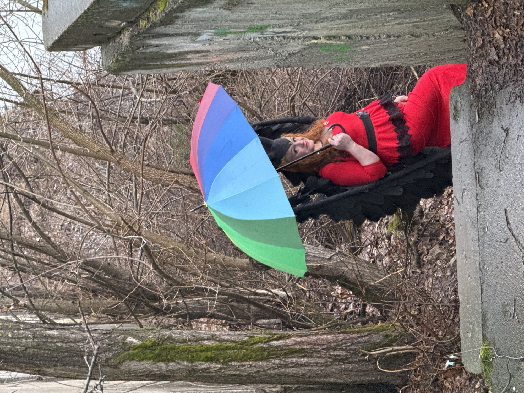 A woman in a red dress and black wings lies in the woods with a colorful umbrella. The location is in the city of Chaikovsky, Russia. The woman is holding the umbrella handle. The environment is a forest with bare trees and some concrete structures.