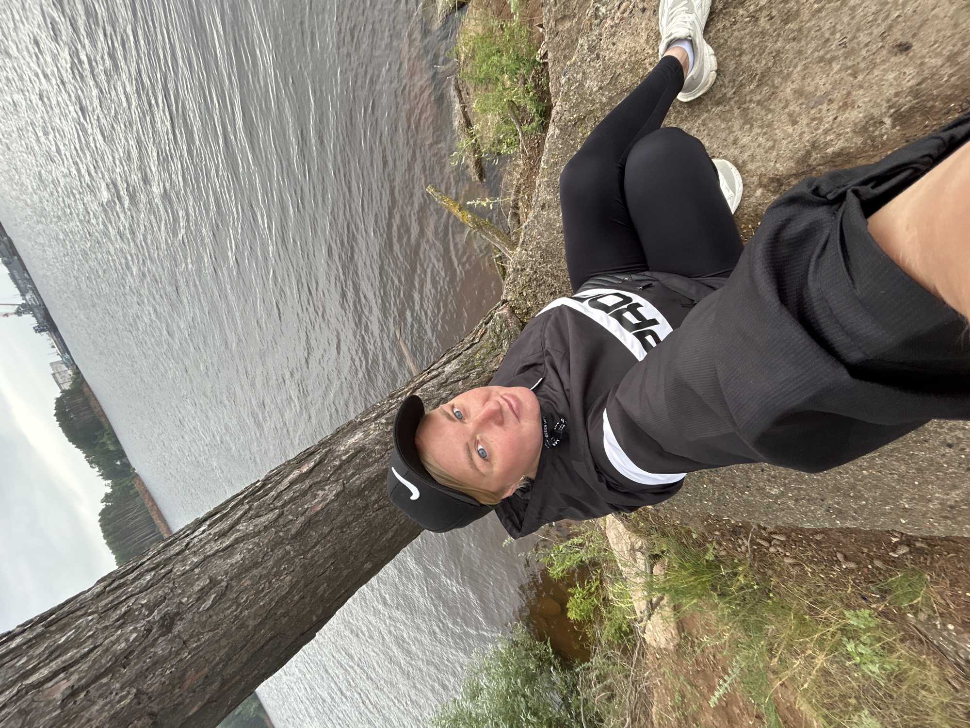 A woman is taking a selfie outdoors, near a body of water. She is wearing a black baseball cap with a white Nike logo, a black jacket with white accents, black leggings, and white athletic shoes. Her face is visible, and she has blonde hair and blue eyes. She is sitting on a concrete surface next to a large tree trunk. The background shows a wide expanse of water, with a treeline on the far side and a cloudy sky. Some buildings and a crane are visible on the far left bank. The scene suggests an overcast day, possibly cool, and a natural or semi-natural outdoor setting. The woman appears to be looking at the camera with a neutral expression. Visible text on her jacket includes "PRO" and "DESIGN".