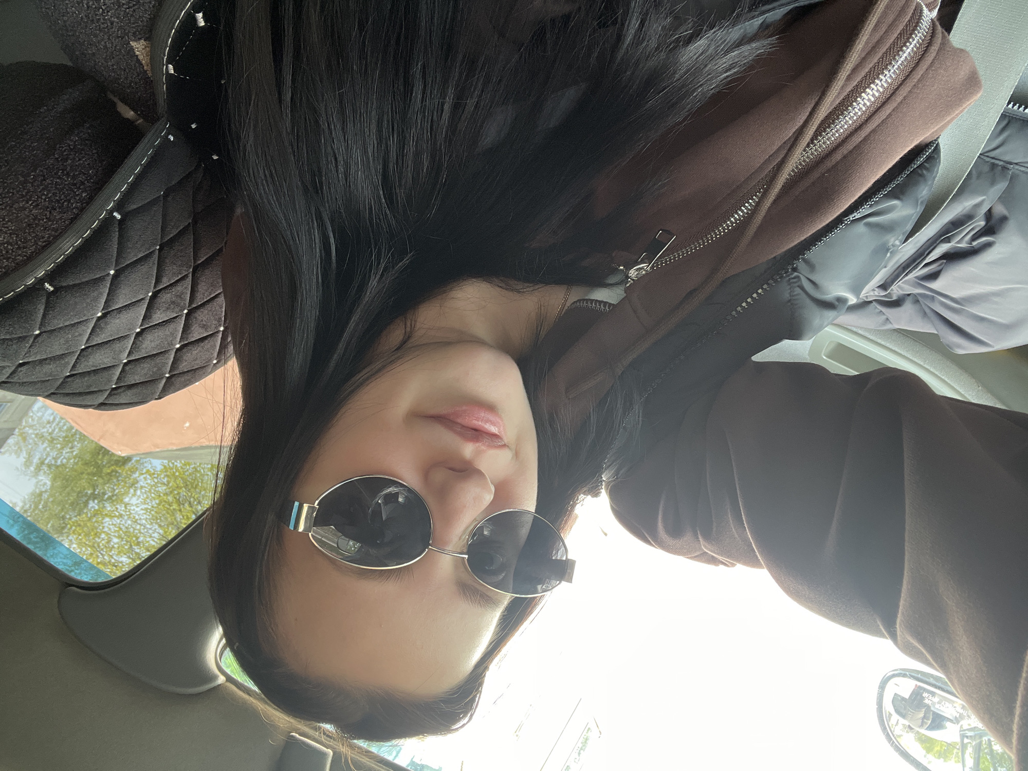 A woman with dark hair is wearing sunglasses. She is inside a car with a black quilted seat cover, wearing a brown jacket. Outside the car, foliage and a building can be seen through the windshield. The location is Chaikovsky, Russia.