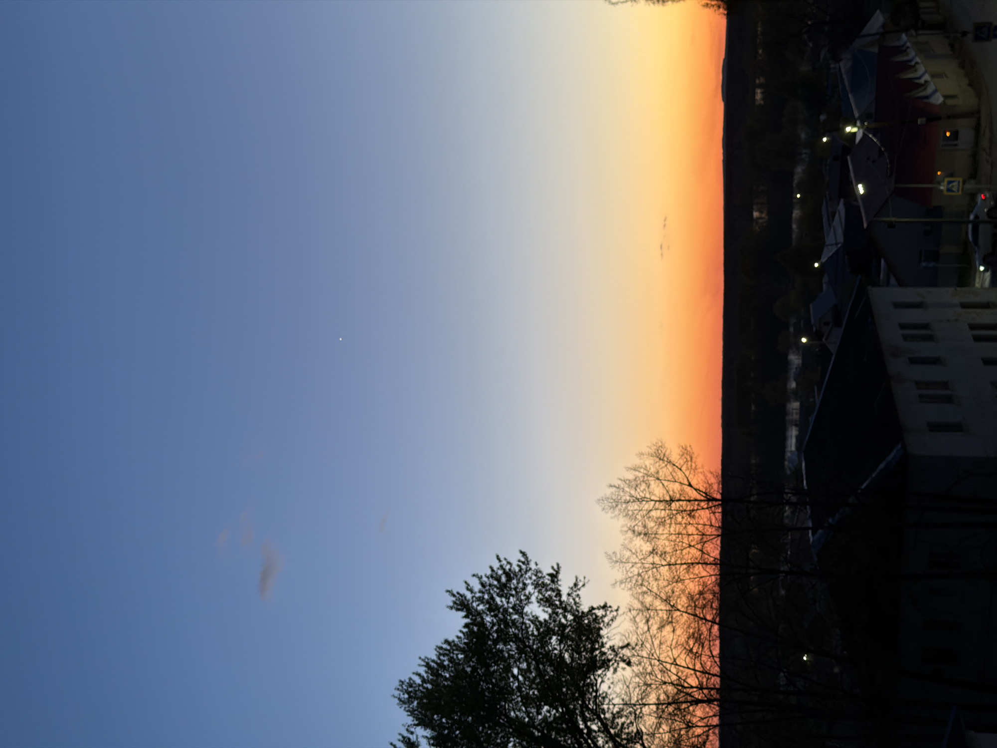 The image depicts a twilight scene in Chaikovsky, Russia. The sky transitions from a deep blue to an orange-tinged horizon. Silhouette trees occupy the lower portion of the frame. City buildings, illuminated by street lights, are visible to the right. A pedestrian crossing sign is barely visible near the buildings. There are no visible people in the frame.
