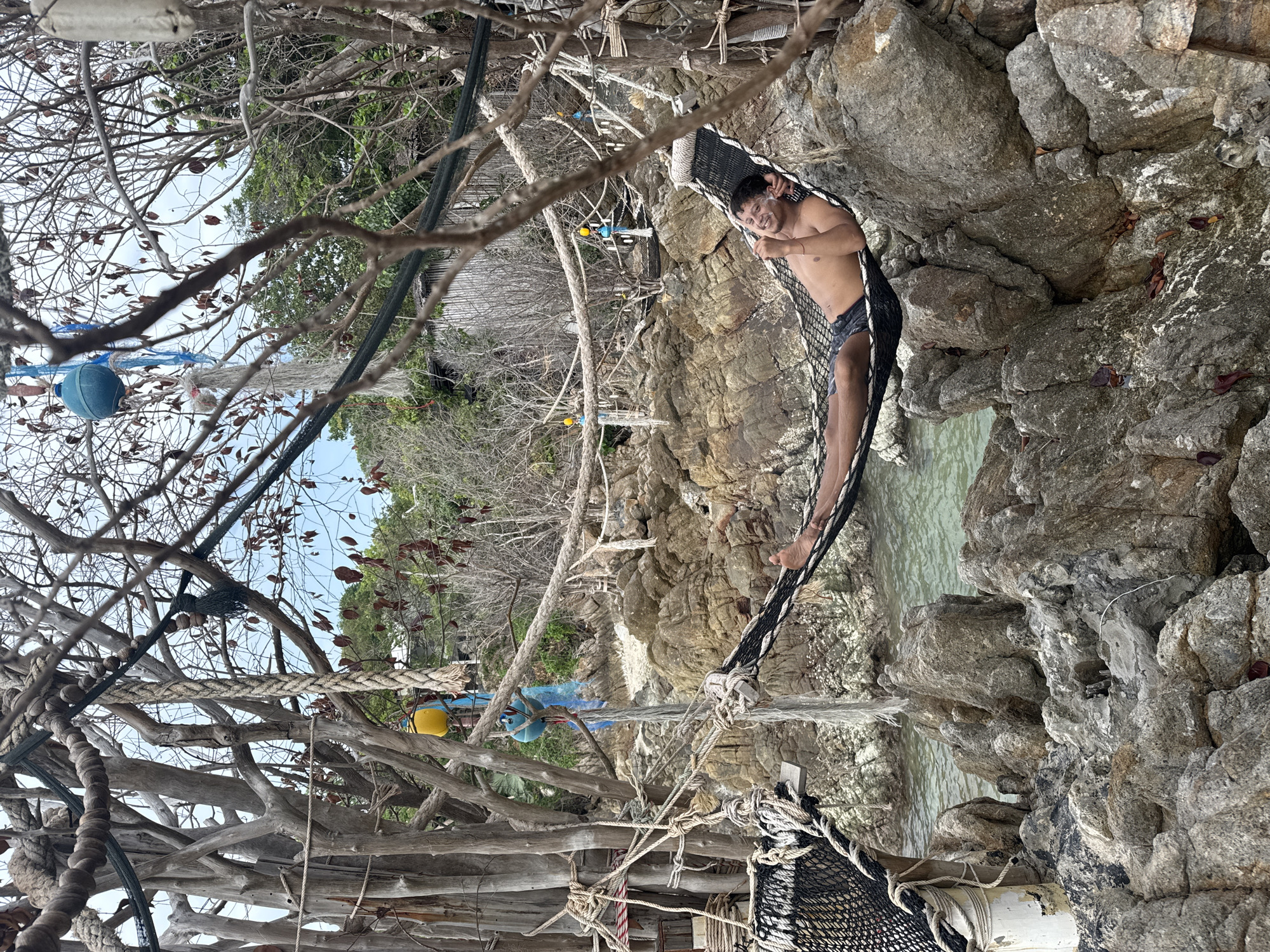 A man reclines in a net hammock strung between rugged rocks on the island of ตำบลเกาะพะงัน, Thailand. The man, shirtless and wearing dark shorts, looks toward the camera with a smile. The hammock is woven from dark material with light-colored accents. The rock formation on the right of the frame is heavily textured and slopes down toward a body of water that shows variations in color, suggesting depth and aquatic life. A complex network of weathered wooden branches and ropes spans the left side of the image. A blue float hangs among the branches. Beyond the wooden structure, there is a glimpse of trees and a light blue sky. The overall composition suggests a serene and secluded beach setting.