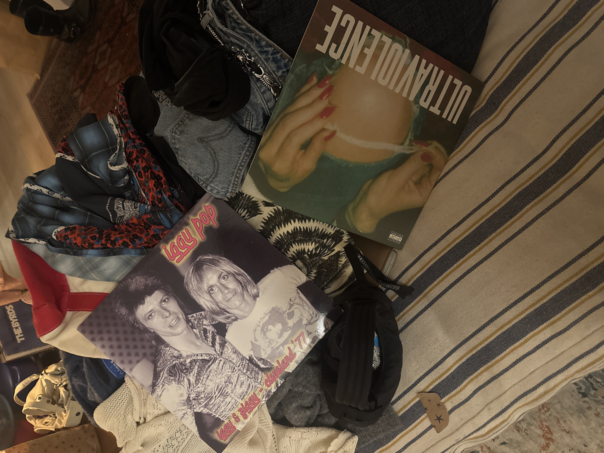 The scene depicts a cluttered indoor space, possibly a living area in the Village of Red Hook, United States of America. Dominating the composition are two vinyl records: one with the title "Ultraviolence" and another titled "Iggy Pop." Various items of clothing are scattered around, including denim jeans, a red and white jacket, and patterned fabrics. A black helmet and a white knitted garment are also present. The objects are arranged on a striped textile or rug. The overall impression is one of disarray and informal storage.