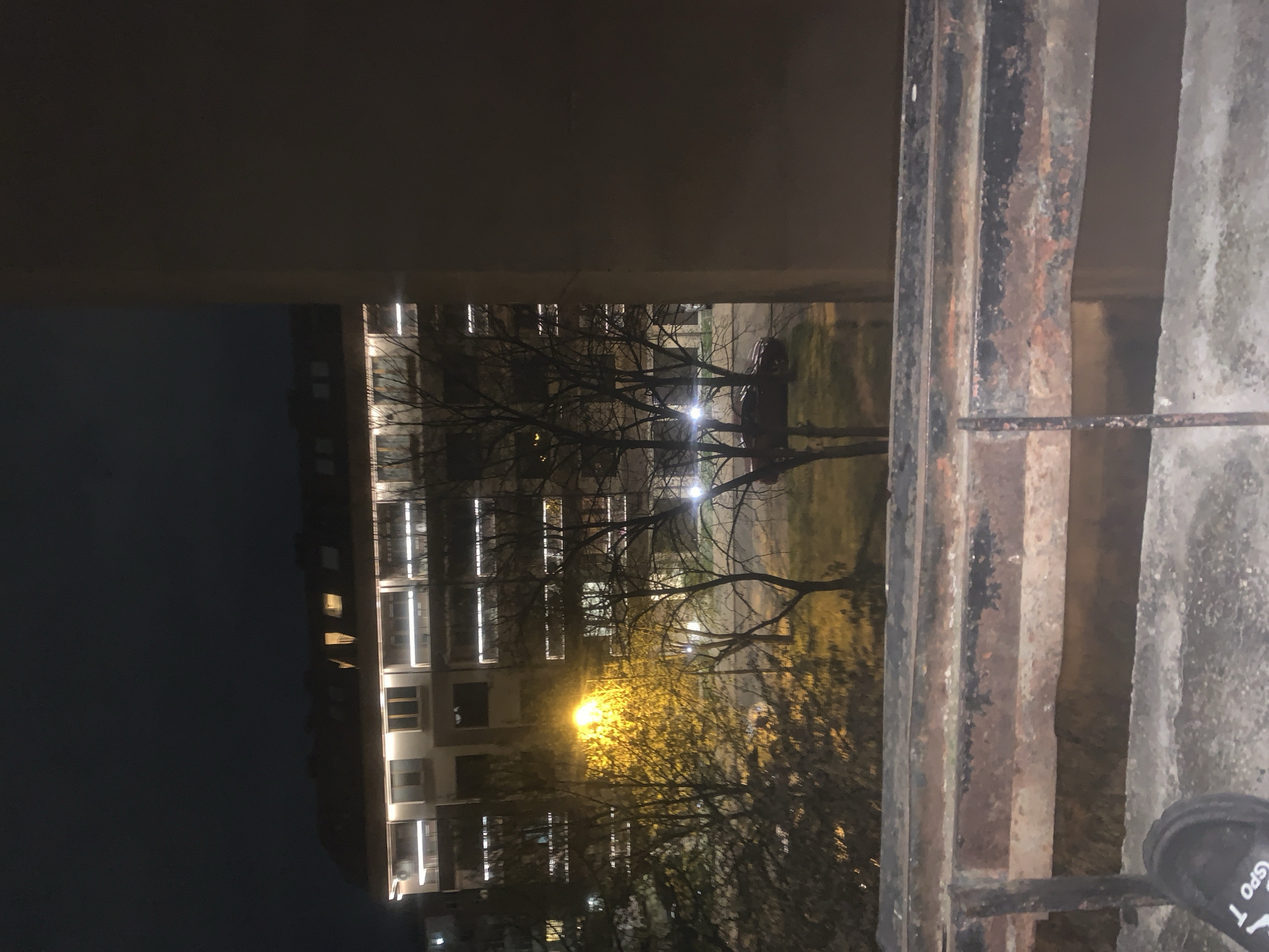 The image is taken from an elevated position, likely a balcony or window, in Sofia, Bulgaria, based on architectural styles and common urban features. The foreground consists of a corroded metal railing. Beyond the railing, a multi-story building dominates the view. Windows are illuminated. Trees are visible between the building and the camera. A street lamp is positioned in front of the building. A dark sky fills the upper left corner. A shoe is visible in the lower-right corner of the frame.