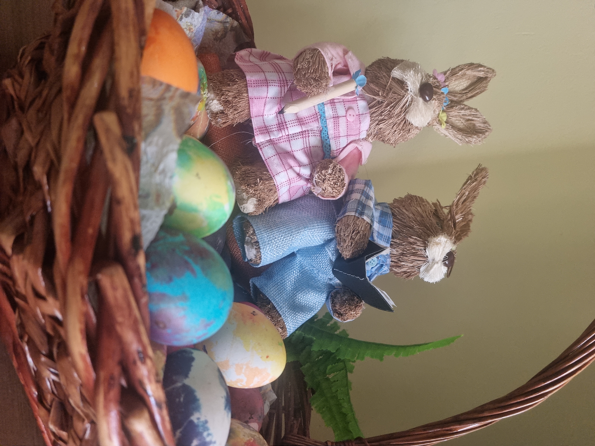 A low-angle shot depicts a wicker basket filled with painted Easter eggs and two straw bunny figurines, likely inside a home. The basket is predominantly brown and composed of woven wooden strips. Several Easter eggs, colored with various shades of orange, yellow, blue, and other mixed tones, fill the basket's left side. The bunny figurines, made of straw, are dressed in plaid shirts – one pink and one blue – and are positioned near the top right of the basket. The straw bunnies have brown eyes and are adorned with decorative flowers. A green fern leaf peeks out from beneath the bunnies. The background is a solid, light yellow color, likely a wall.