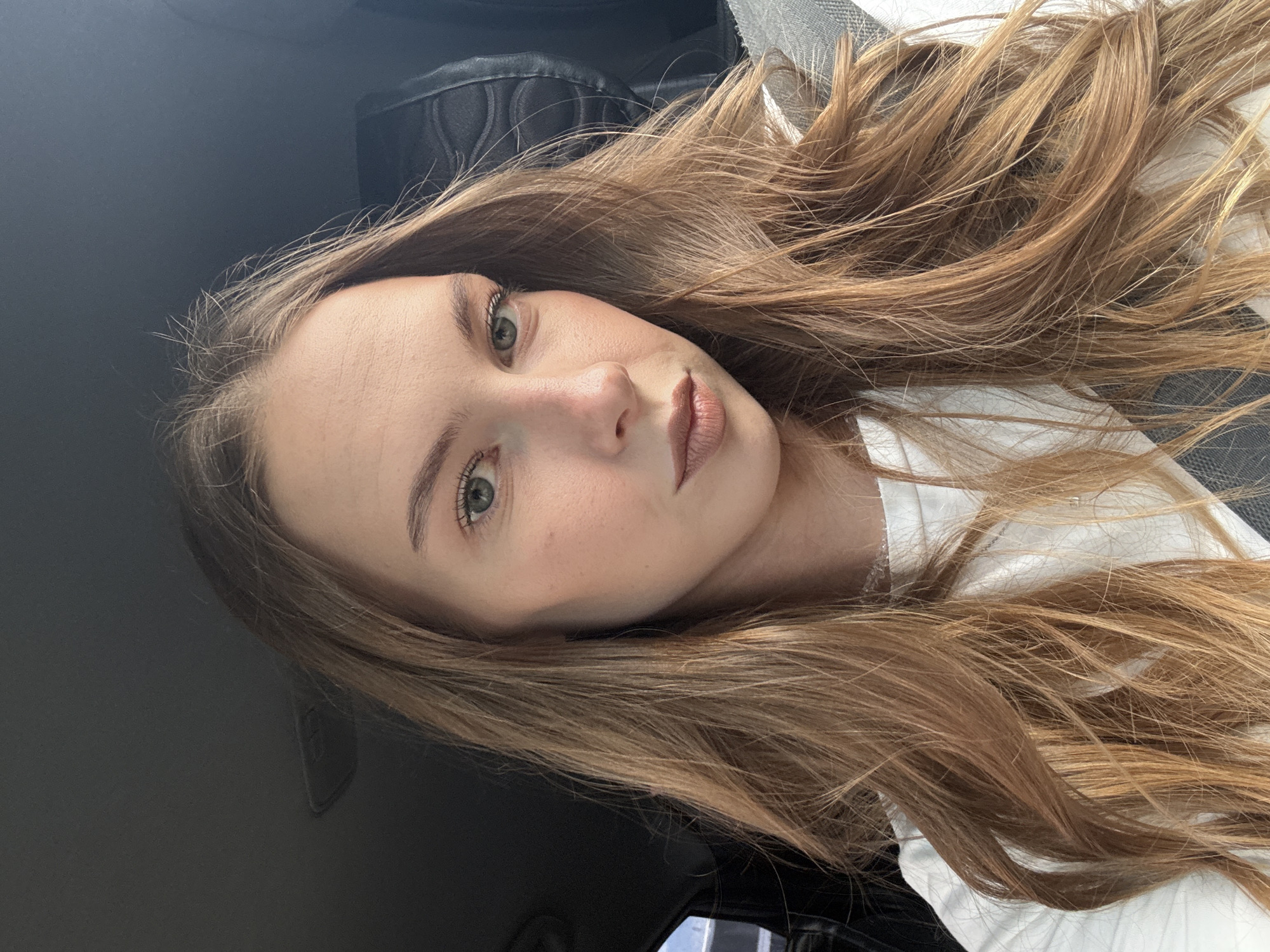 A young woman with long, wavy, light brown hair and green eyes is pictured in a selfie taken from the chest up. She is wearing a white t-shirt and is seated in what appears to be the driver's seat of a car. The background shows a car's interior, including a dark-colored headrest and a grey seatbelt. The image is a close-up, focusing on the woman's face and hair. The location is not specified in the image or its context as being in Perm, Russia.