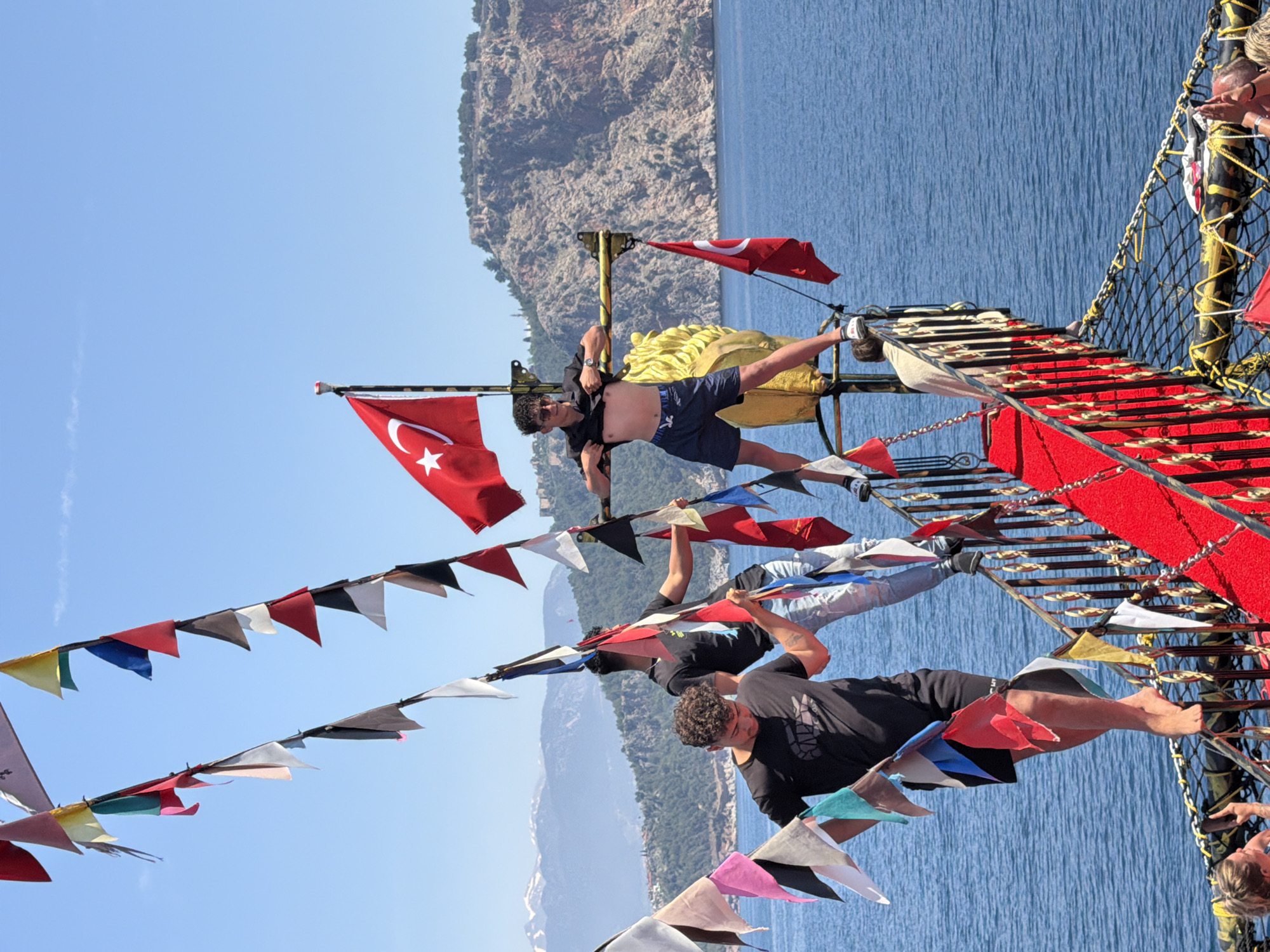 The image shows a sunny day on a boat near the coast of Alanya, Turkey. The blue sea stretches out, with mountains visible in the background. On the boat, two men hang from ropes decorated with colorful triangular flags, apparently posing for photos. One man is bare-chested and wearing shorts and sunglasses. Turkish flags are also visible. The boat has a red carpet on a higher platform, and people are on the deck or climbing up the sides.
