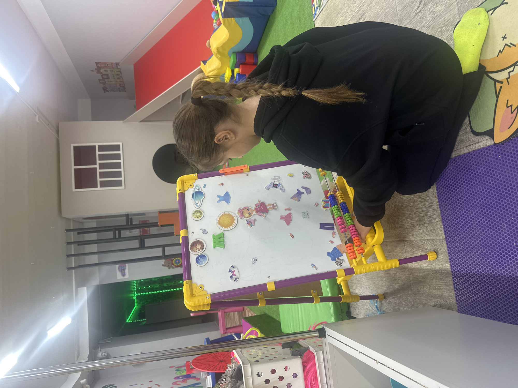 An individual, likely a child or young adult, with light brown hair styled in a braided ponytail, kneels on a light wooden floor. They are wearing a black hooded sweatshirt and vibrant yellow-green socks, and are actively engaged with a colorful whiteboard easel. The easel features a yellow and purple frame and is covered with various magnets, including cartoon characters, suns, planets, clothing items, and a distinct green character or symbol. An abacus-like toy with colorful beads is attached to the side of the easel.
The setting appears to be an indoor play or learning area, characterized by a multi-textured floor with sections of light wood, green carpet, and a purple honeycomb-patterned mat. In the background, soft play equipment in blue, yellow, and green hues is visible. The room's walls are a mix of white and light gray, with a prominent red accent wall. A rectangular window with a grid design is present on one wall, and further back, a green laser light grid is projected onto another surface. A small sticker depicting colorful houses adorns the ceiling. Additional elements include white shelving units and a metal rack holding various items, including a bright pink wig and a tiara. The space is brightly lit, suggesting an indoor environment with artificial lighting. The person's focus on the easel indicates an activity of play or learning.