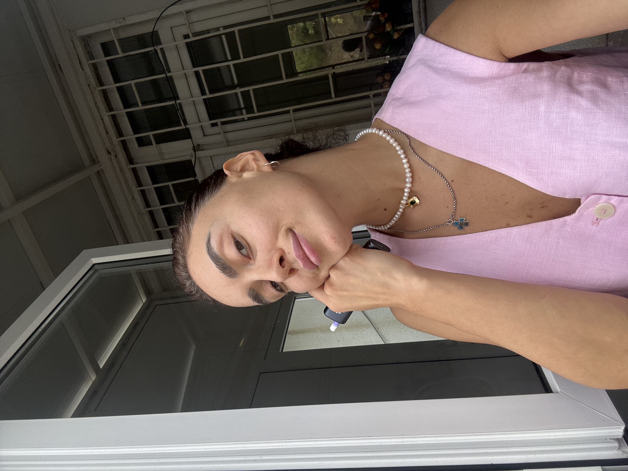 A young woman with dark hair pulled back is posing for a selfie outdoors. She is leaning her head on her hand, holding a small black electronic device in her fingers. She is wearing a pink sleeveless v-neck top and two layered necklaces, one with pearls and a gold charm, the other with a silver chain and a blue and silver cross pendant. She has a subtle smile and is looking directly at the camera. The background shows a white window frame on the left and a barred window with a white exterior frame on the right. The lighting suggests it is daytime. The overall scene appears to be a residential balcony or window overlooking an urban environment, consistent with a location in Tbilisi, Georgia, where such architectural styles are common.