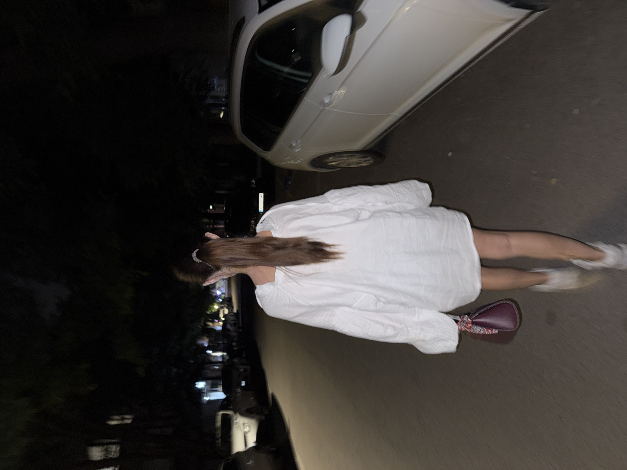 The image captures a nighttime scene on a street in Plovdiv, Bulgaria, showing a person from behind, standing or walking next to a parked white car. The photo is rotated 90 degrees clockwise, depicting the individual vertically on the right side.
The main subject is a person with long, straight brown hair, styled with a light-colored hair clip. They are wearing a loose-fitting, long-sleeved white dress that extends to mid-thigh. White sneakers are visible on their feet, with some motion blur suggesting movement. A small, burgundy-colored handbag with a patterned strap is slung over their shoulder, resting against their hip.
To the left of the person, a white car is parked on what appears to be a paved road or sidewalk. Only the side and part of the front bumper and wheel are visible. The setting is dark, indicating it is night. In the background, to the left of the car and the person, there's a dark area that appears to be dense foliage or trees, with faint, distant artificial lights hinting at other buildings or street activity further down the road. The street surface is a dark grey, with some subtle variations in texture. There is no visible text in the image.