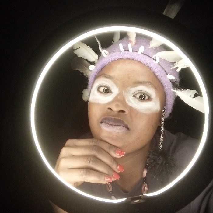 A person is centered within a ring light's illumination. The person wears a lavender beanie adorned with white feathers and small white beads. White makeup streaks emphasize the eyes. The person's hand is raised to their chin. They are wearing a shirt that appears to be lavender and a necklace made up of oval beads. The ring light contrasts with the dark background surrounding the person.
