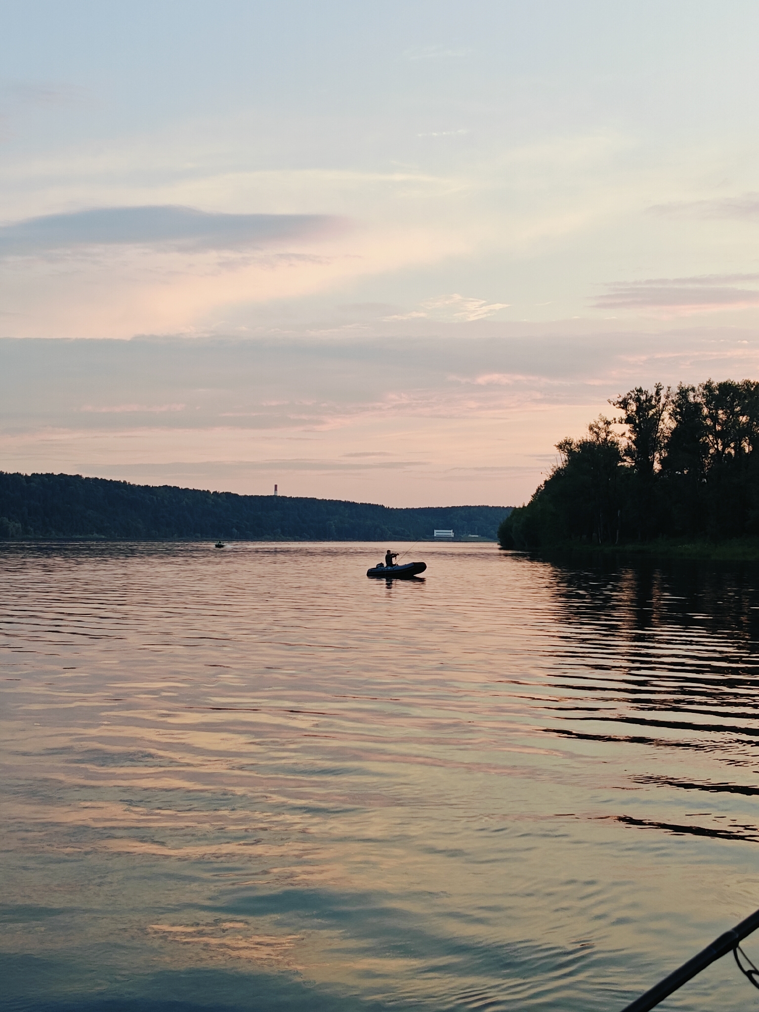 The image depicts a tranquil sunset scene on a wide river in Noviy, Russia. The sky is painted with soft hues of pink, orange, and pale blue, with wispy clouds scattered across it. The water's surface reflects the colors of the sky, creating a shimmering, rippling effect. In the middle of the river, a small inflatable boat with a single occupant, silhouetted against the fading light, is visible. The figure appears to be fishing, holding a rod. The riverbanks are lined with dense, dark green trees, creating a stark contrast with the colorful sky and water. In the distance, along the far bank, a faint outline of buildings and a red tower can be discerned. The overall atmosphere is peaceful and serene, capturing a moment of quiet solitude by the water.