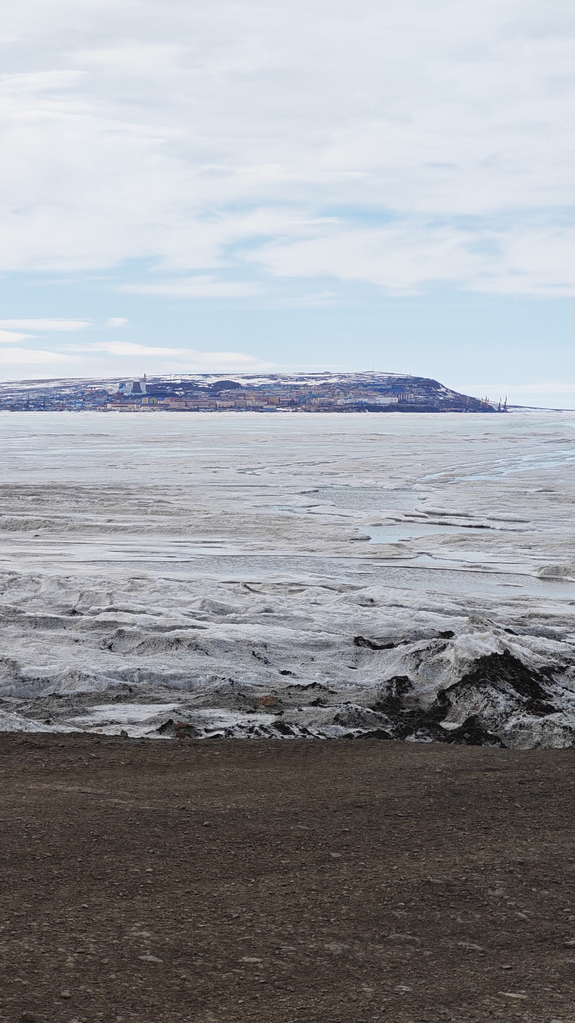 The image depicts a landscape with a city skyline in the background, likely in Ugolnye Kopi, Russia. The foreground consists of a dark, coarse ground. Above it, a large expanse of icy or snowy terrain stretches towards the middle ground, marked by rough textures, ridges, and some pooling water. In the distance, a cluster of buildings and structures form a city skyline against a cloudy sky. The color palette is dominated by browns, grays, and whites, with touches of blue in the sky.