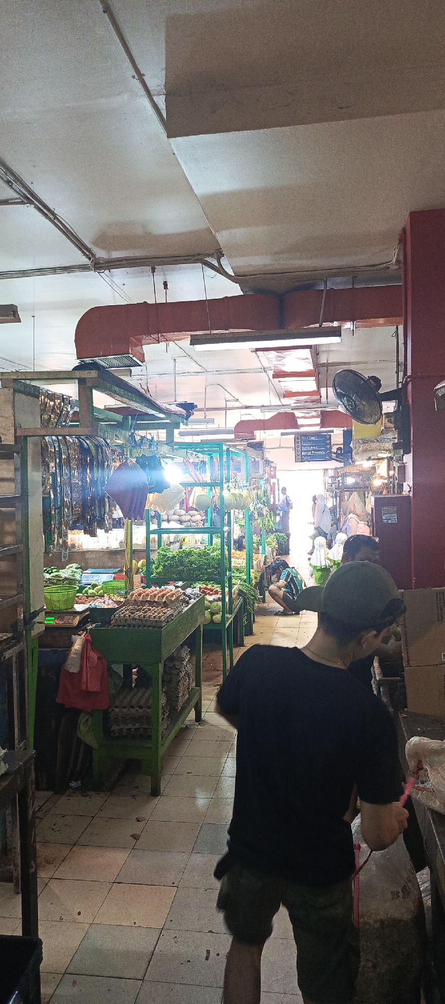 The image depicts an indoor marketplace, possibly a wet market, in Sukaresmi, Indonesia. In the foreground, a person wearing a black t-shirt and olive green shorts is seen from the back, with their head turned away from the camera. They appear to be interacting with something off-camera to their right, possibly a stall or a seller. The marketplace is filled with various stalls displaying fresh produce, including vegetables and what appear to be eggs. The stalls are constructed with green metal frames, and some have wooden tops. Overhead, industrial-style ventilation ducts and lighting fixtures are visible, suggesting a functional, perhaps older, facility. Several other individuals are present in the background, some appearing to be shoppers or vendors. The floor is tiled, and the overall impression is one of a busy, functional marketplace.