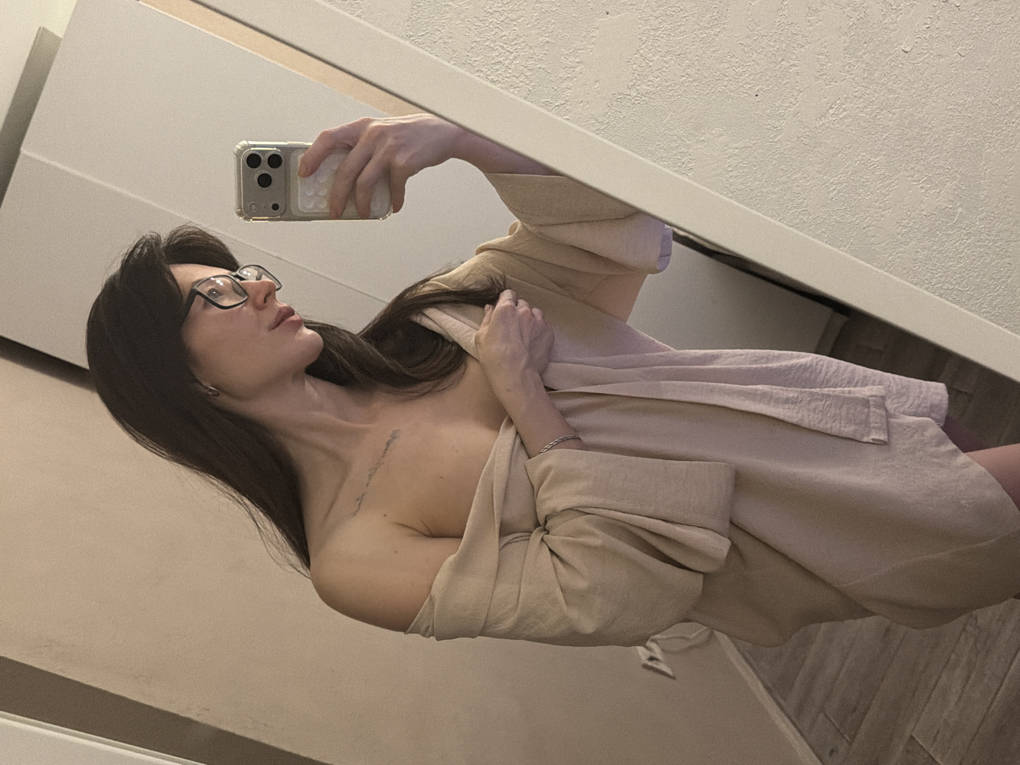 A woman with long dark hair and glasses is taking a selfie in a mirror. She is wearing a light-colored robe or wrap that is partially open, revealing her upper chest and shoulder. She holds a smartphone in her right hand, with the camera facing the mirror. The background shows elements of a room, including a textured wall and what appears to be a cabinet or dresser. The location of Perm, Russia is not discernible from this image.
