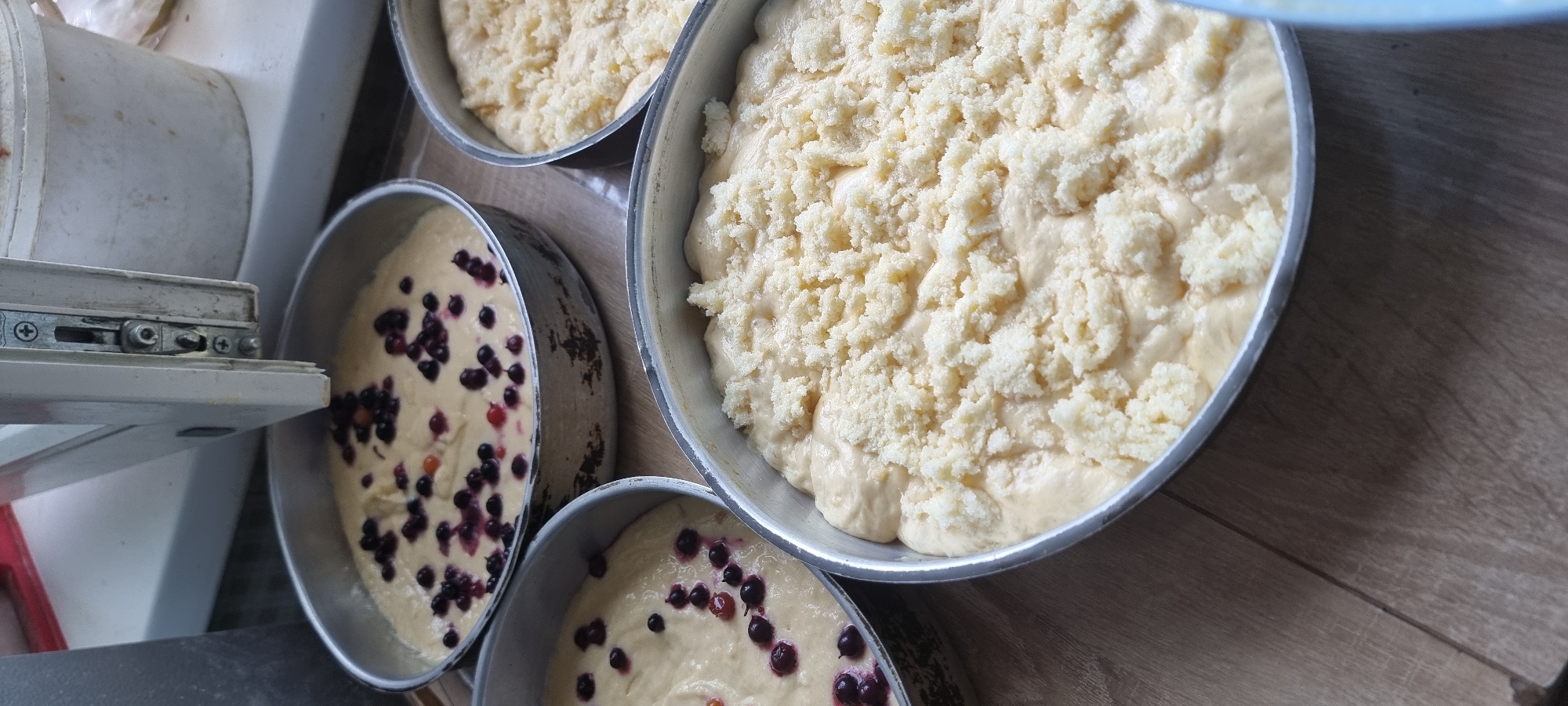 Four round metal baking pans are arranged on a wooden surface. Three of the pans are partially visible. The largest pan, in the foreground and slightly to the right, is filled with dough and topped with a crumble topping. To its left and slightly behind is another pan containing dough with dark berries mixed in. Behind that, to the left and further back, is a third pan with dough and dark berries. A fourth pan is visible to the far right, also appearing to contain dough with a crumble topping. In the background, a white bucket and a portion of a white object with metal fixtures are visible. The lighting suggests an indoor setting. The image provides no information regarding the city of Aktobe, Kazakhstan, or any people.