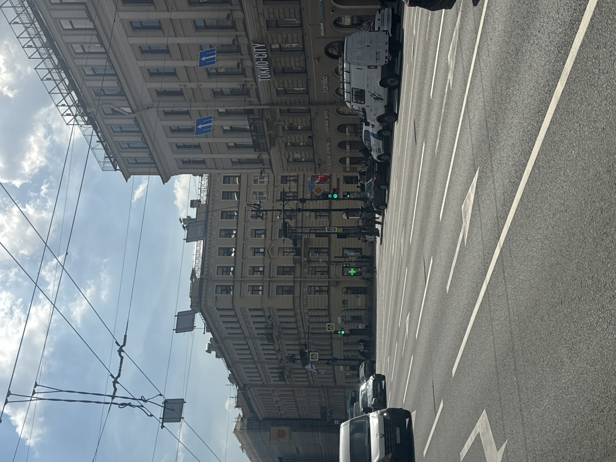 The image depicts a wide street in Saint Petersburg, Russia, on a cloudy day. Several buildings with multiple floors line the right side of the street, featuring numerous windows and architectural details. Traffic lights and street signs are visible. Several vehicles, including vans and cars, are on the road. Pedestrians are seen on the sidewalks. Overhead, a complex network of wires, likely for public transportation like trams, crisscrosses the sky. The road surface is asphalt with clear lane markings.