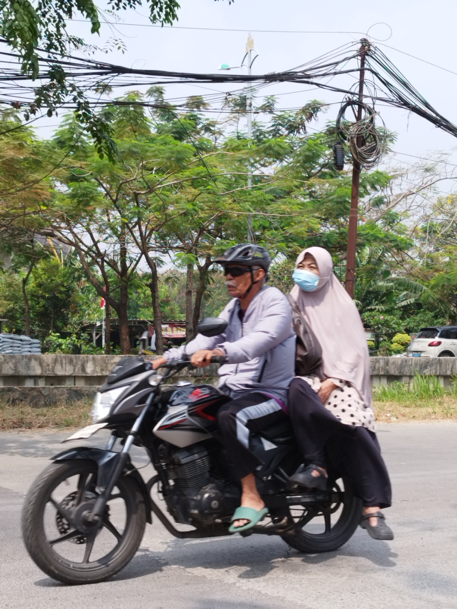 A middle-aged man wearing a helmet and sunglasses, and a woman wearing a hijab and a face mask are riding on a motorcycle on a paved road in Bekasi, Indonesia. The man is driving the motorcycle, and the woman is sitting behind him. Both are dressed in casual clothing. The motorcycle is black and silver, with red accents. The background shows a row of trees and some overhead power lines. There is a grey concrete barrier along the side of the road, with some bags of material stacked behind it. A white car is visible in the distance. The sky is overcast.