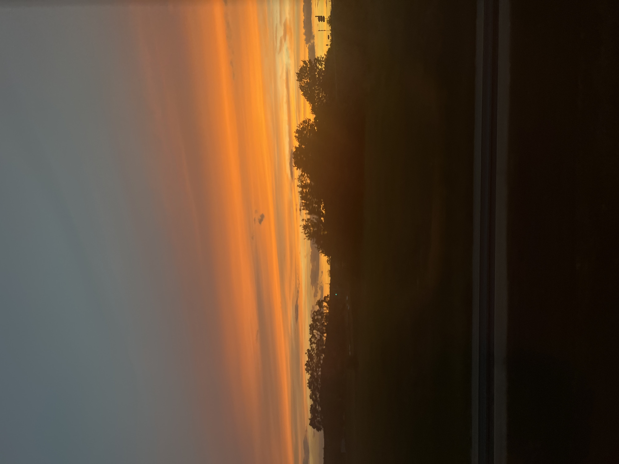 The image shows a sunset over a rural landscape with a silhouetted treeline. The sky is filled with vibrant colors of orange, yellow, and a hint of blue towards the top, indicating the transition from day to night. A dark foreground suggests a field or body of water, with a distinct horizontal line composed of two dark gray bands possibly representing a railing or a fence. The general atmosphere is serene and natural. No people or specific man-made structures are clearly identifiable in Baiersdorf, Germany, due to the silhouetting and the focus on the natural elements.