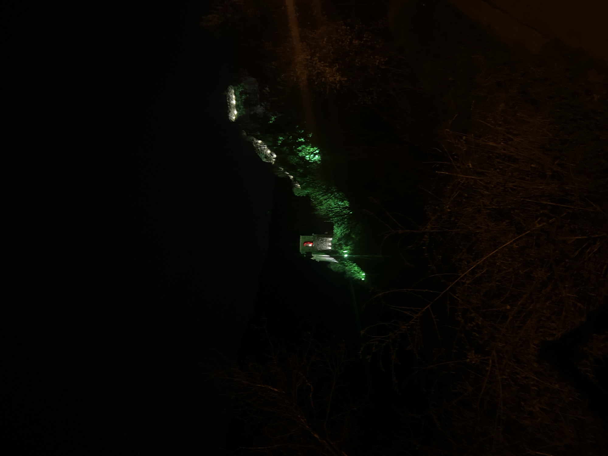 At night in Asenovgrad, Bulgaria, a brightly lit building sits along a walking path. The structure is at the end of the path. The environment is heavily forested. The path is lit with green lights.