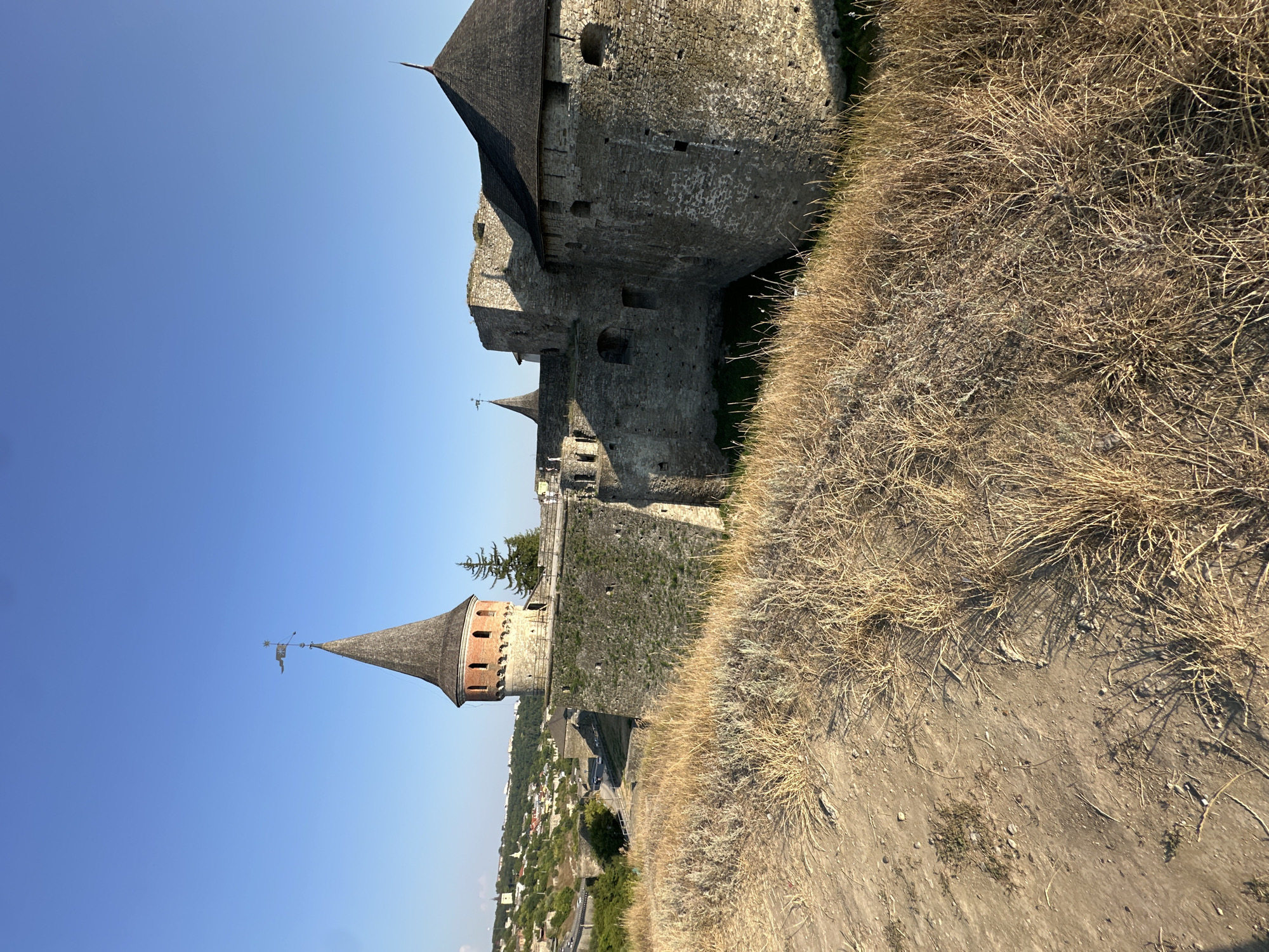 The image shows a view of the Kamianets-Podilskyi Castle in Kamianets-Podilskyi, Ukraine. The castle is a well-preserved fortress with stone walls and conical towers. The foreground is dominated by dry, yellow grass and a dirt path, suggesting a dry, possibly late summer or early autumn season. In the background, the castle's stone walls and a prominent conical tower with a flag are visible under a clear blue sky. A section of the city, with buildings and trees, can be seen on a hill to the left of the castle. The sun is shining, casting shadows on the dry grass and the castle walls. No people are visible in the image.