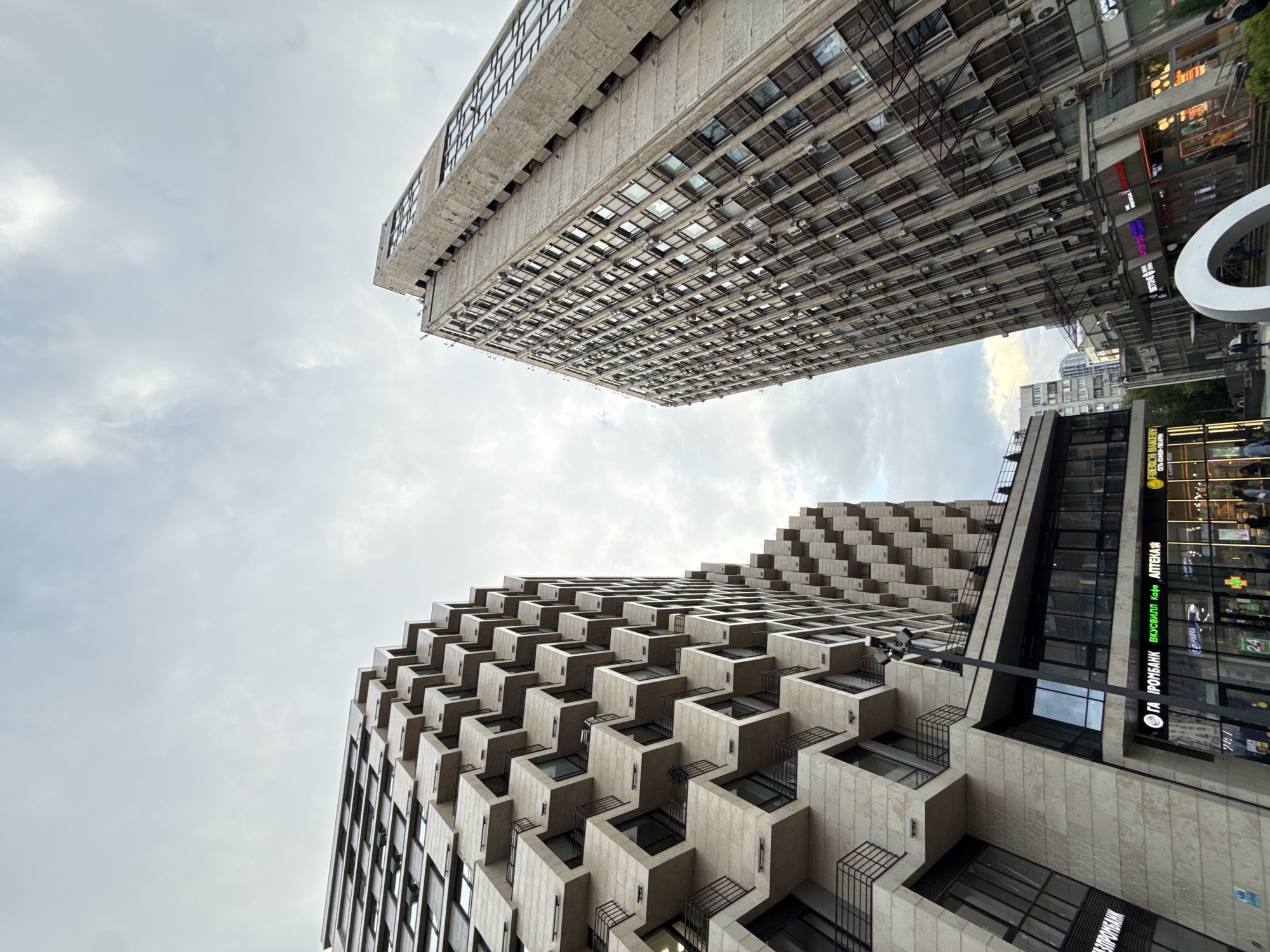 The image shows an upward view of two buildings in Moscow, Russia, set against a cloudy sky. The building in the foreground has a distinctive stepped façade, with each level projecting outward, forming individual balconies or terraces. Dark window frames contrast with the light-colored stonework. A section of the building to the right transitions into a more conventional façade with large windows displaying interior activity, signage, and storefronts. Text in both Cyrillic and Latin alphabets is visible, including the phrases "ГА ІРОМБАНК", "ВКУСВИЛЛ Кафе АПТЕКА", and "FRIENICH BAKERY". A street light is visible, connecting the architecture with the ground.
In the background, the second building is a taller, rectangular structure with a repetitive grid pattern of windows. The building exhibits a boxy design with horizontal rows of windows and air conditioning units.