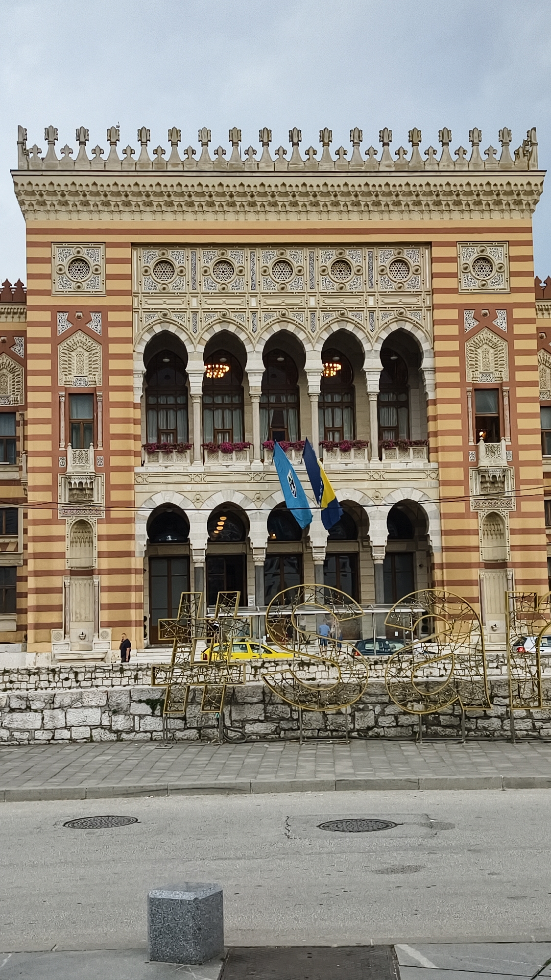 The image shows the Sarajevo City Hall, also known as Vijećnica, a prominent building in Sarajevo, Bosnia and Herzegovina. The building features a distinctive Austro-Hungarian architectural style with Neo-Moorish elements, characterized by its striped facade of red and yellow sandstone, numerous arched windows, and ornate detailing. Two flags are displayed on poles in front of the building: a blue and white flag with a crest and a yellow and blue flag.
In the foreground, a stone wall serves as a separation between the building's entrance area and the street. Behind this wall, several people are visible, including a man standing near the wall and others walking or standing in the background. A yellow taxi is parked behind the wall, and other vehicles are present further down the street. A large, intricate golden metal structure, possibly an art installation or part of a festive decoration, is positioned in front of the stone wall, partially obscuring the view of the building's lower level. The street in front of the installation is paved with asphalt and features manhole covers. A small, grey granite cube is placed on the pavement to the left of the image. The sky above is overcast, indicating a cloudy day.
