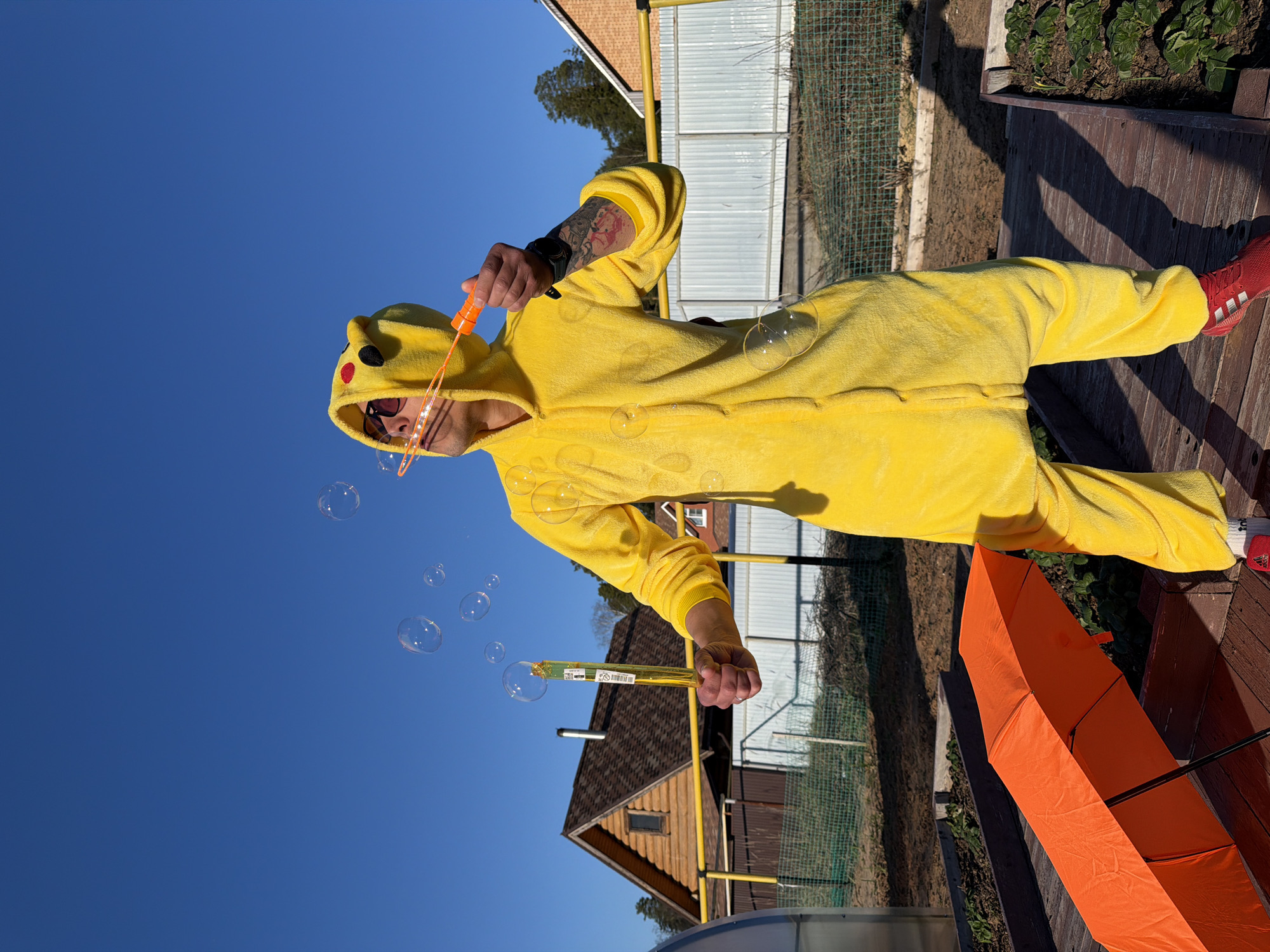 A person in a Pikachu onesie is blowing bubbles outside on a sunny day in the city of Chaikovsky, Russia. The person is wearing sunglasses and red shoes. They are standing on a wooden deck with an orange umbrella nearby. The background includes a clear blue sky, a house, and a fence.