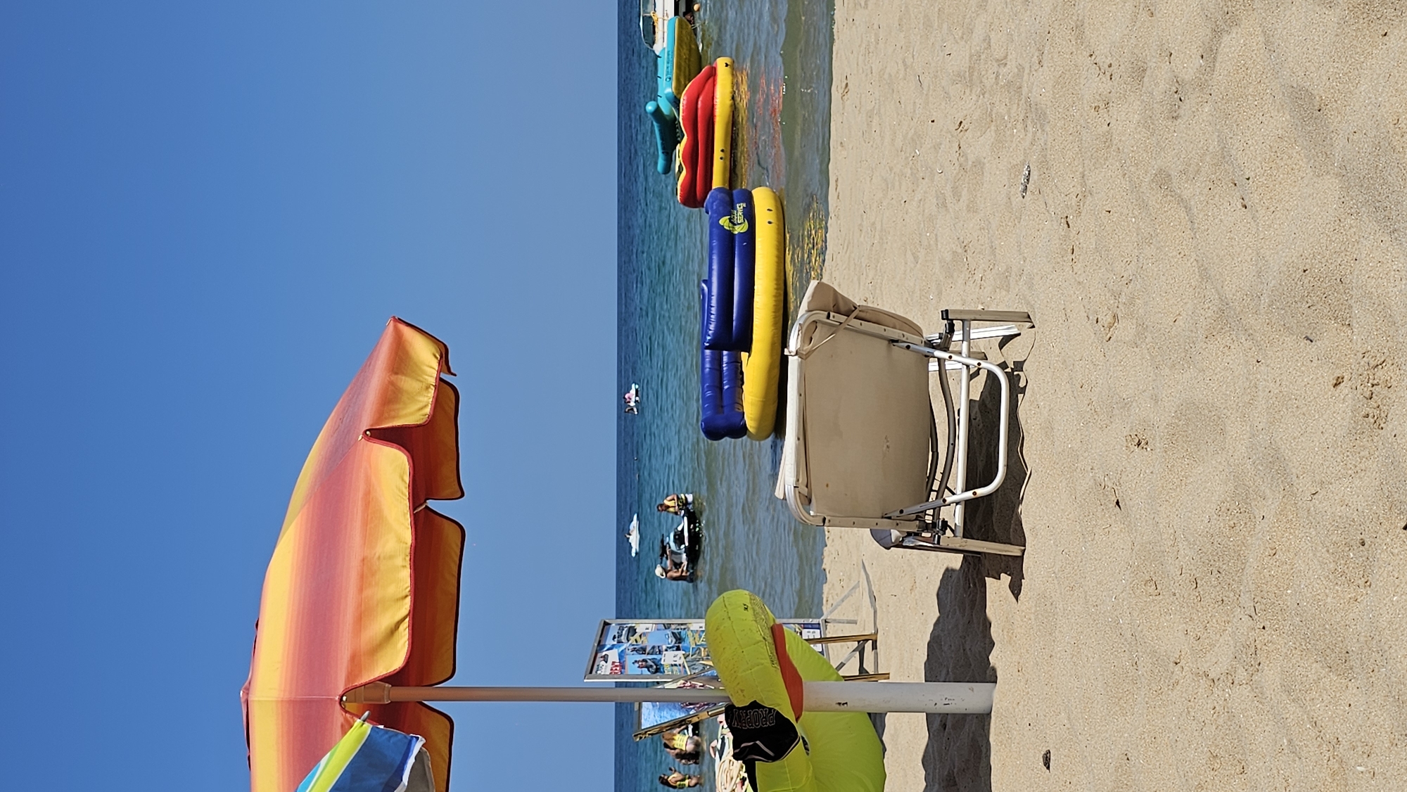 The image depicts a sunny day at a beach, likely in Varna, Bulgaria, given the context. In the foreground, a sandy beach stretches out, with a white lounge chair positioned towards the center. To the left, a colorful beach umbrella shades a part of the sand. Beyond the beach, the tranquil blue sea is visible. Several inflatable water toys, including a yellow and blue one and a red and yellow one, are floating on the water. Further out, people are seen enjoying various water activities, with jet skis and other small boats on the surface. The sky is a clear, bright blue. The scene evokes a relaxed atmosphere typical of a summer beach day.