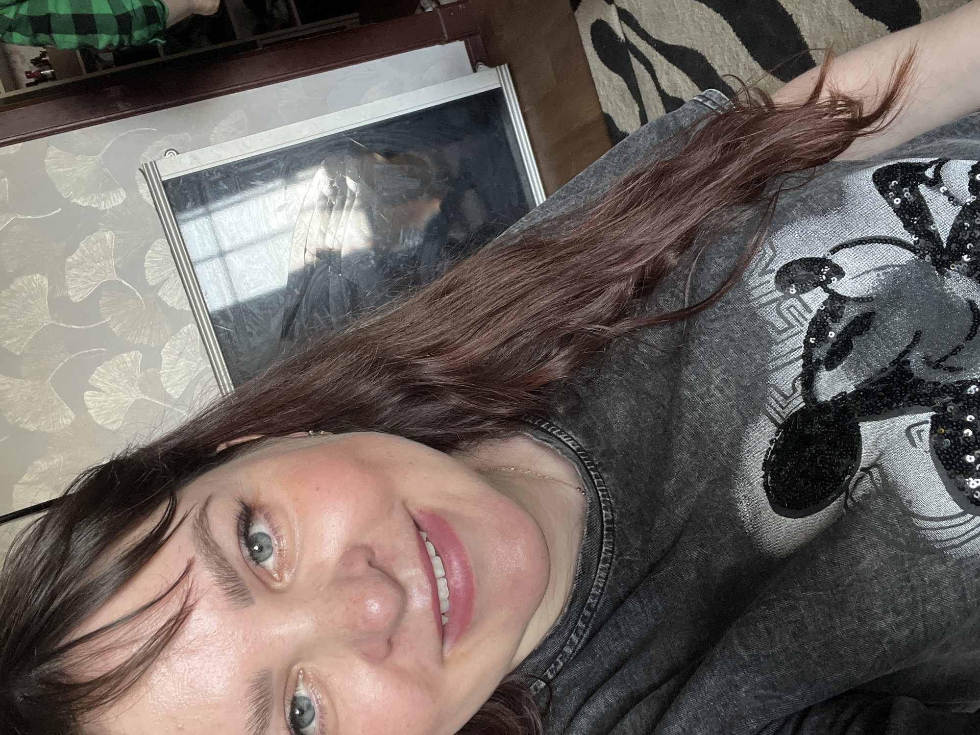 A selfie features a woman with light skin, brown hair, and blue eyes. She is smiling. She wears a grey t-shirt. Behind her is a wall with a ginkgo leaf pattern, a framed mirror reflecting a person in a green and black plaid shirt, and a zebra-striped surface. There is no evidence to confirm the location is Chaikovsky, Russia.
