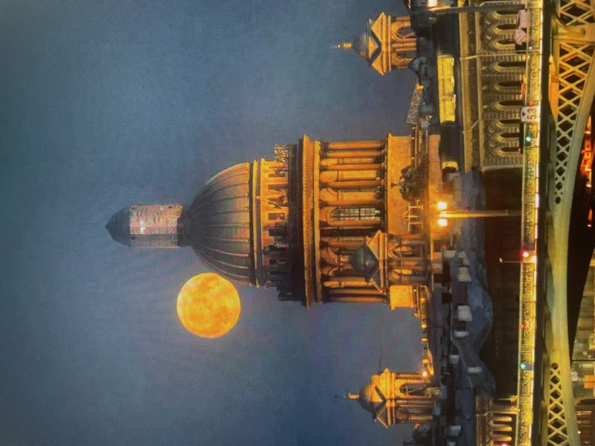 The image depicts an orange moon rising behind a large, domed building with illuminated windows. The building appears to be an older, grand structure, possibly a cathedral or government building, with ornate architectural details. To the right of the building, a bridge with illuminated arches and crisscrossed supports is visible. The sky is a deep, dusky blue, suggesting it is either dusk or dawn. The overall scene evokes a sense of wonder and perhaps a touch of mystery, with the luminous moon and the majestic architecture. The bridge and building suggest an urban setting, likely near a body of water, though no water is directly visible. The image appears to be a photograph, possibly taken at a distance to capture both the moon and the architectural elements. No people are visible in the image.