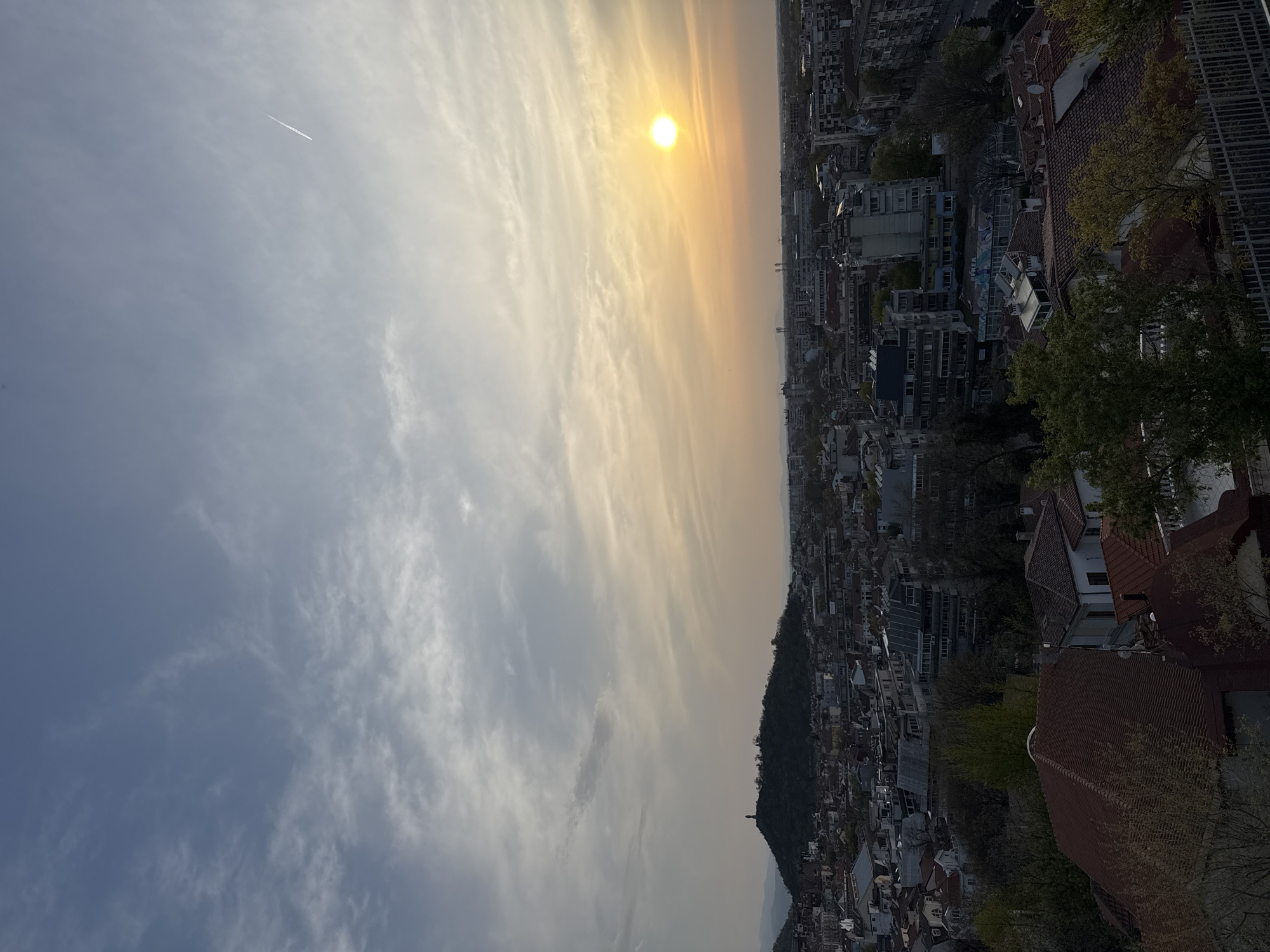 An aerial daytime shot, taken at a tilted angle, shows the city of Plovdiv, Bulgaria, at sunset. The sky is mostly cloudy. A contrail can be seen in the top left of the sky. The buildings below are packed together in a tight formation. The foreground shows roof tops, trees, and a metal railing. The sun can be seen setting over the city.