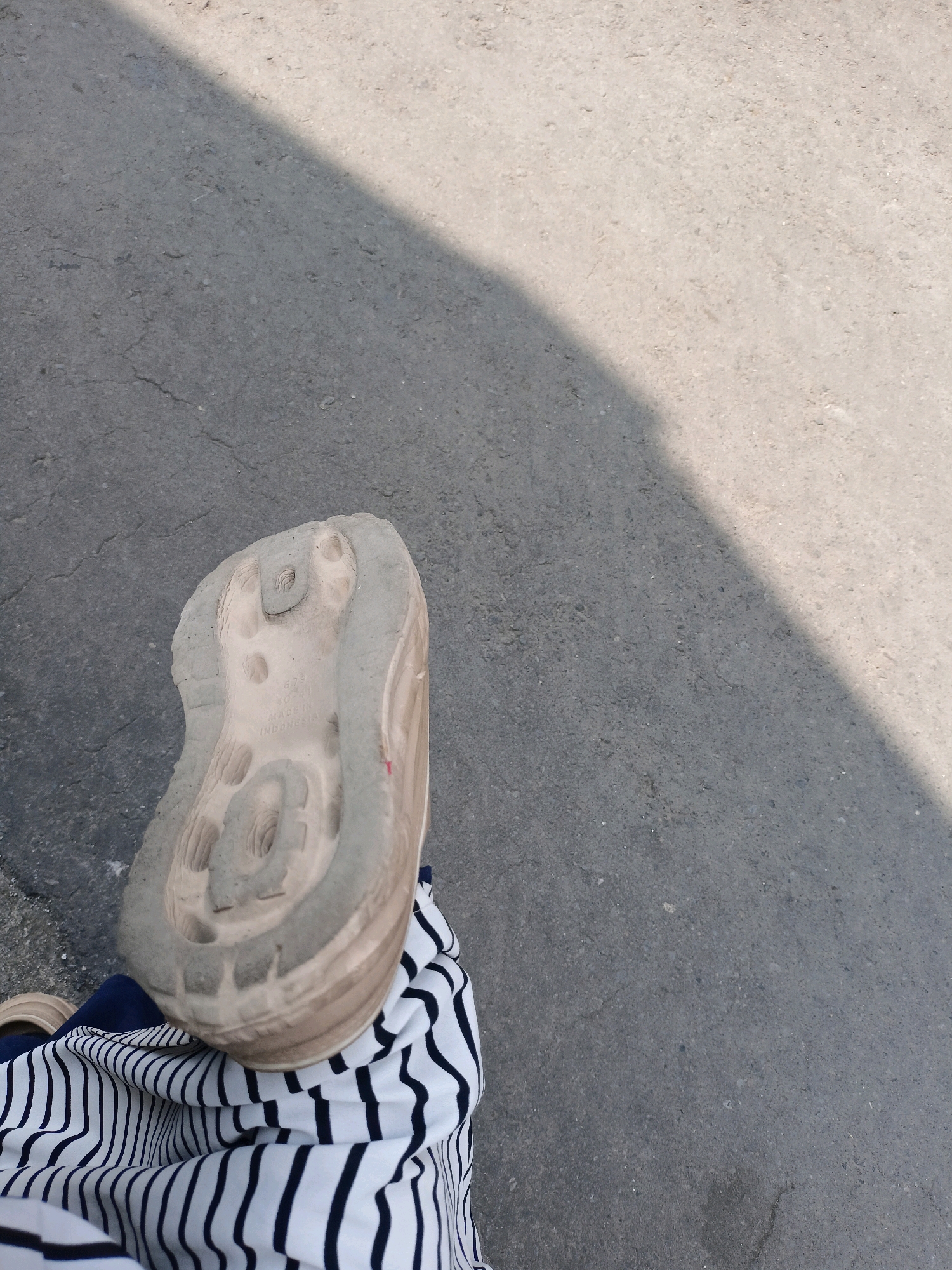 The image shows the sole of a dirty, light-colored shoe resting on a gray asphalt surface. A shadow from an unseen object falls diagonally across the asphalt. The shoe appears to be a sneaker, with visible treads and some text embossed on the sole, including "MADE IN INDONESIA". The person wearing the shoe is also wearing striped pants, with black and white stripes. The setting appears to be an outdoor urban environment, consistent with the city of Payaman, Indonesia. There are no other people or significant objects visible in the frame.