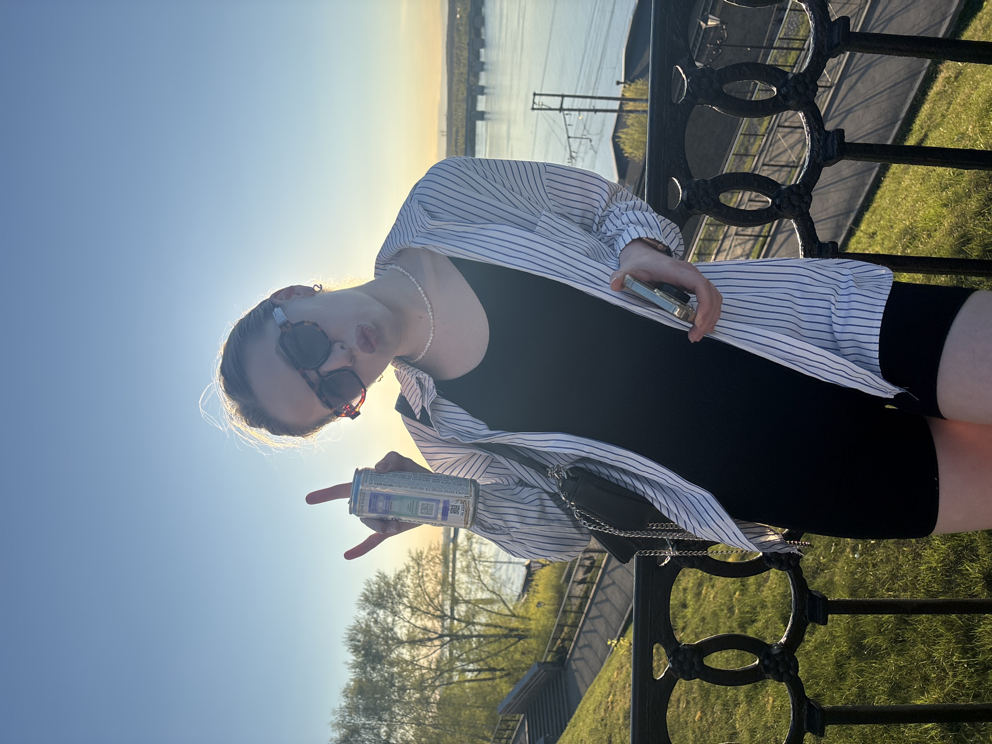 A young woman stands outdoors by a black wrought-iron fence. She wears sunglasses, a pearl necklace, a black dress, and a white and blue striped shirt. She holds a can of "Fraga Dolce" drink with a view of the Kama river in the background. The scene is located on the Embankment in the city of Perm, Russia.