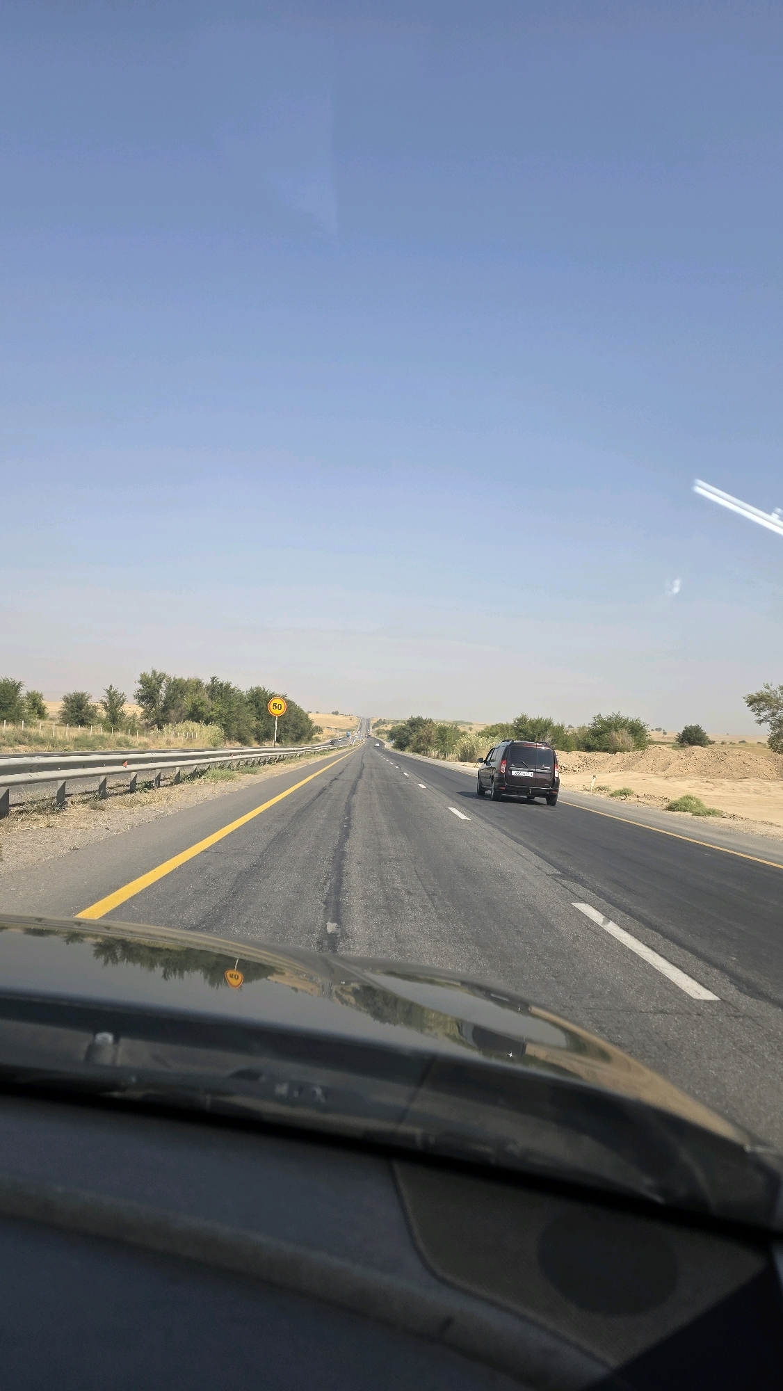 The image depicts a view from inside a vehicle, looking forward along a multi-lane highway. The road is paved and marked with yellow and white lines. Trees and sparse vegetation line the sides of the highway, with a dusty, arid landscape extending beyond. A speed limit sign indicating "50" is visible on the left side of the road. Two dark-colored vehicles are present on the highway, one in the left lane and another in the right lane ahead. The sky is clear and blue. The foreground shows the interior of the vehicle, including the dashboard and windshield, reflecting the surrounding environment.