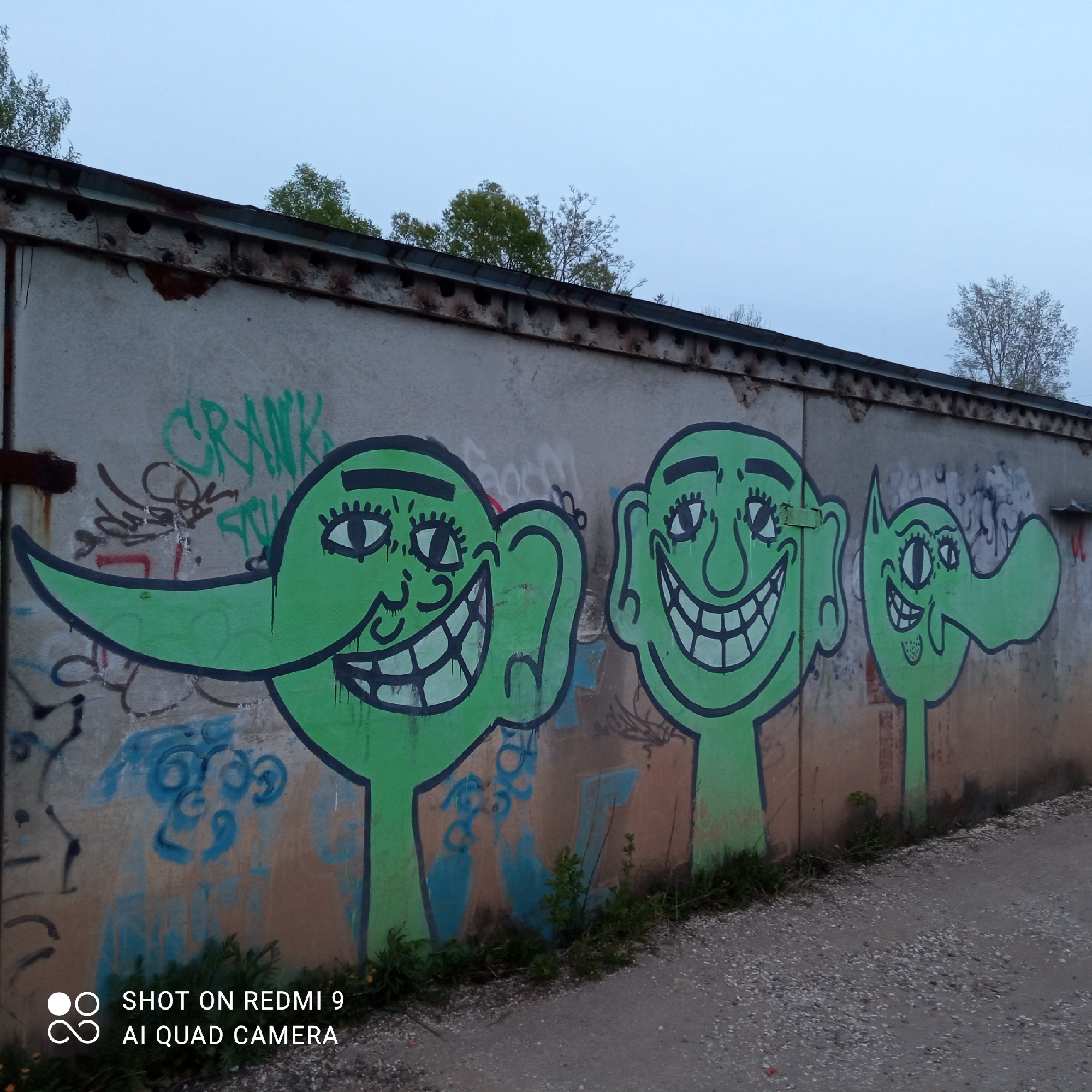 The image shows graffiti art on a weathered concrete wall, likely a part of an industrial or utilitarian structure, possibly a garage or utility building in Chaikovsky, Russia. The graffiti depicts three cartoonish green figures with exaggerated facial features, large eyes, and prominent smiles. The wall also has other layers of older graffiti. The foreground shows a gravel path or road. The sky is overcast, suggesting evening or early morning light. Small weeds and plants are growing at the base of the wall.