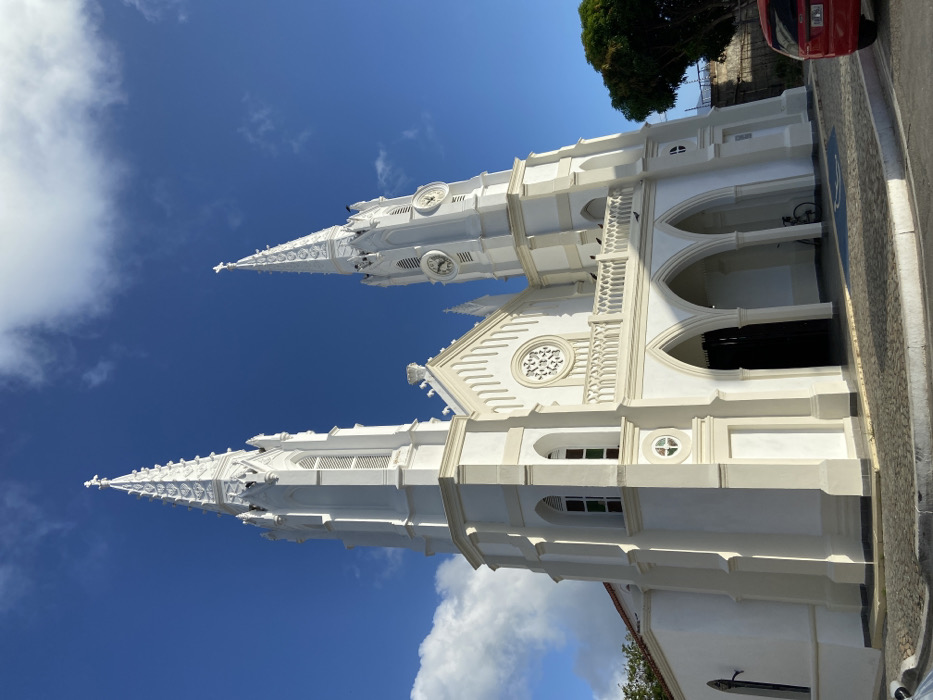 A low-angle shot captures the facade of a striking white church against a vibrant blue sky dotted with fluffy clouds. The church features two prominent towers, each adorned with intricate details and a clock. A red car is visible on the right side of the frame. The architectural style appears ornate, with arched entryways.