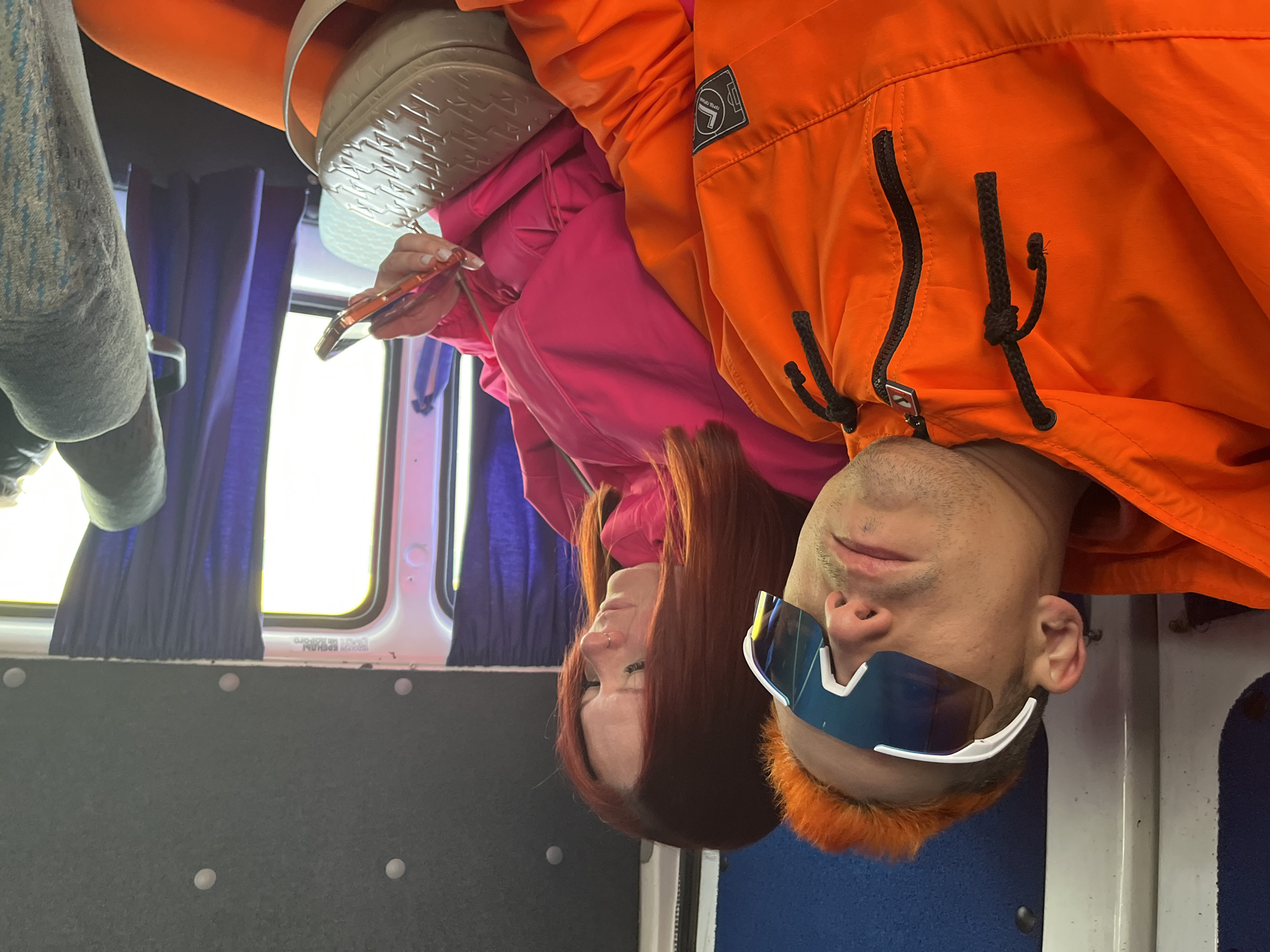 The image shows a man and a woman inside a vehicle, possibly a bus, as suggested by the window and interior paneling. The man has bright orange hair and is wearing sunglasses and an orange jacket. The woman has long, reddish-brown hair and is wearing a pink hoodie. She is holding a phone. The setting appears to be a bus or van interior with blue curtains and grey paneling. The image does not allow for the determination of its location in the city of Chaikovsky, Russia.