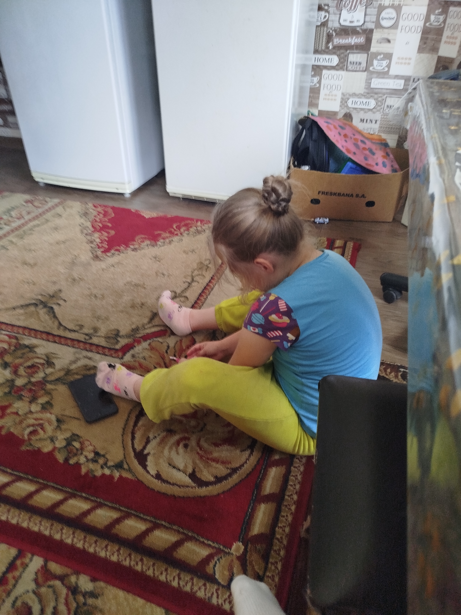 A young girl with blonde hair in a bun sits on a patterned rug in what appears to be a domestic setting. She is wearing a blue t-shirt with colorful patterned sleeves and yellow pants. Her feet are adorned with pink socks with small patterns. In front of her, on the rug, is a dark rectangular object, possibly a phone. The background features two white refrigerators, a cardboard box filled with items including a pink blanket, and a wall with decorative paper depicting coffee-related imagery and text. A portion of a dark, possibly upholstered, chair is visible in the lower right corner of the image. The environment suggests an indoor residential space in Крывасельскі сельскі Савет, Belarus.