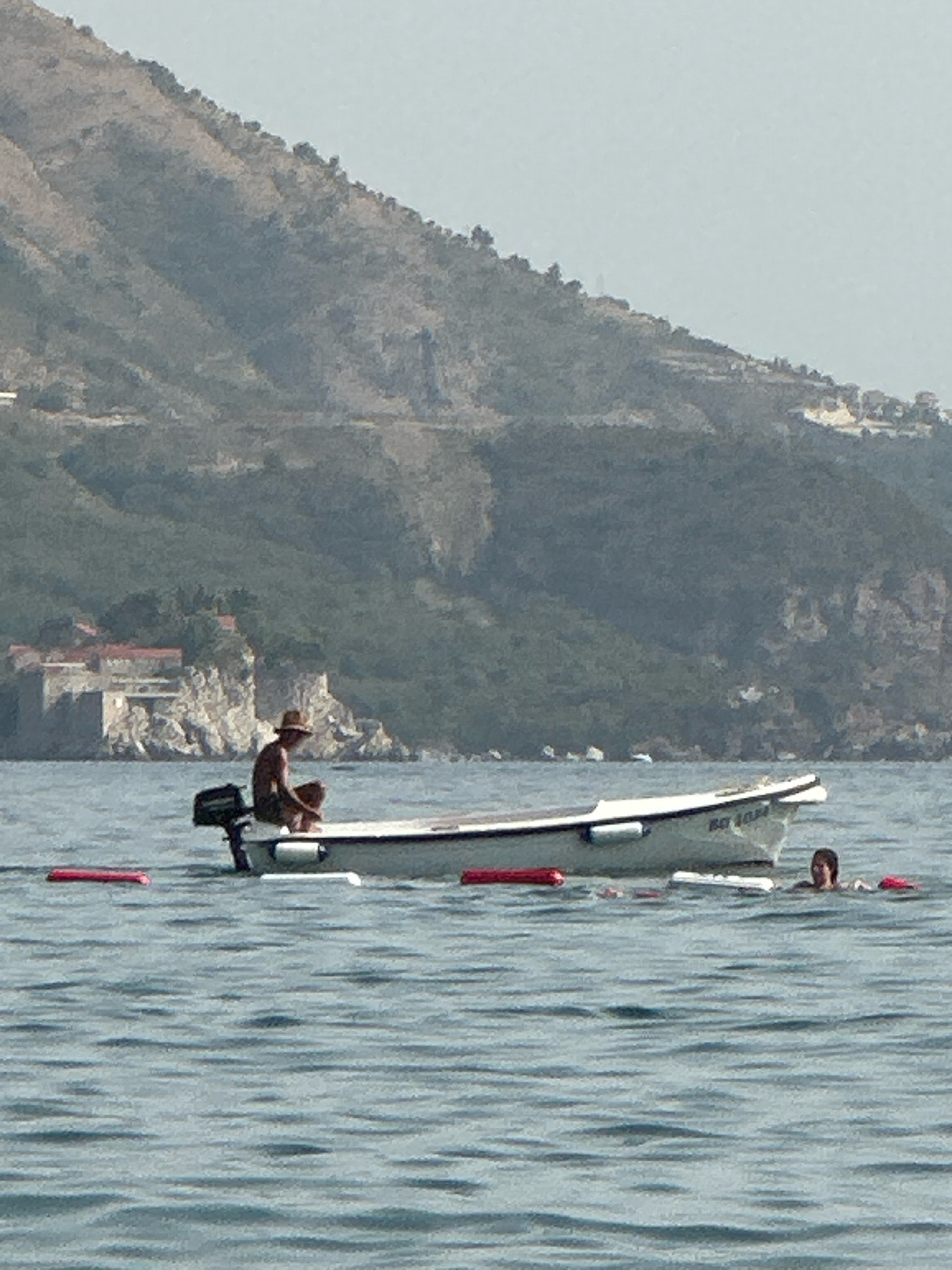 In the waters off the coast of Boreti, Montenegro, a small white boat with an outboard motor is anchored. A person wearing a hat is seated in the boat, appearing to rest. To the right of the boat, two individuals are swimming in the sea, with one of them partially submerged. In the background, a lush green hillside rises steeply, with a rocky outcrop featuring stone buildings and red-tiled roofs. The water is calm, with gentle ripples reflecting the overcast sky. Red buoys are visible floating on the surface, marking a boundary or area. The scene suggests a leisurely day at the seaside, with people enjoying the water and the scenic views.