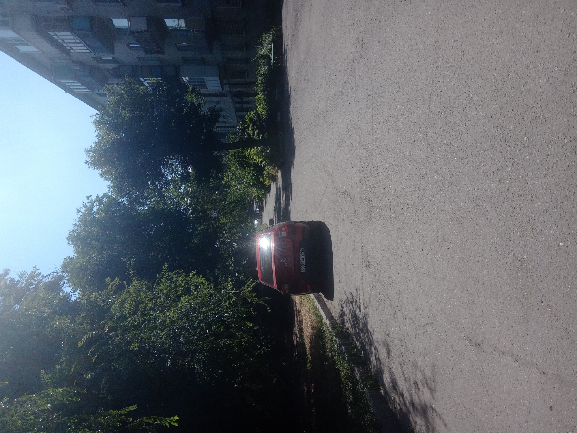 The image shows a street scene in Oskemen, Kazakhstan. A red sedan is parked on the right side of the asphalt road, facing away from the camera. The car has a license plate that reads "891AHY 16". Sunlight glares off the car's rear window. To the left of the road is a sidewalk with a curb, and beyond that, dense green trees and foliage. In the background, on the right side of the image, is a multi-story apartment building with balconies. The sky is clear and blue. There are no people visible in the image.