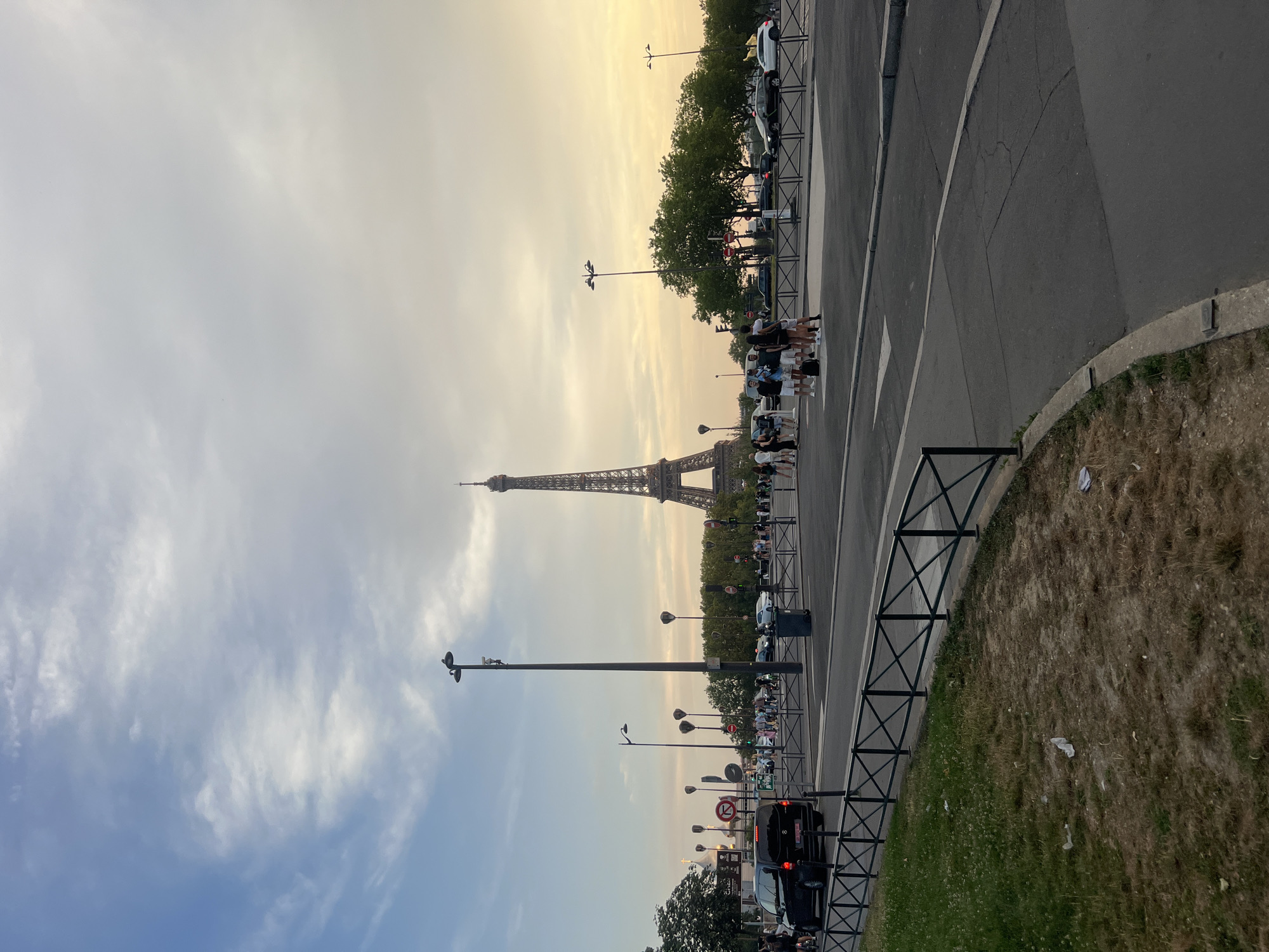 This image captures a view of the Eiffel Tower in Paris, France, during what appears to be the late afternoon or early evening. The sky is a mix of blue and warm, hazy tones, with scattered clouds. In the foreground, a street with a crosswalk is visible, and several cars are on the road, including a black van and some white vehicles. A group of people are standing on the sidewalk or crossing the street. Lampposts are scattered throughout the scene, and trees line the background. The grass area in the bottom left shows some discarded pieces of litter. The iconic Eiffel Tower is prominently featured in the middle-right of the image, towering over the surrounding trees and buildings.