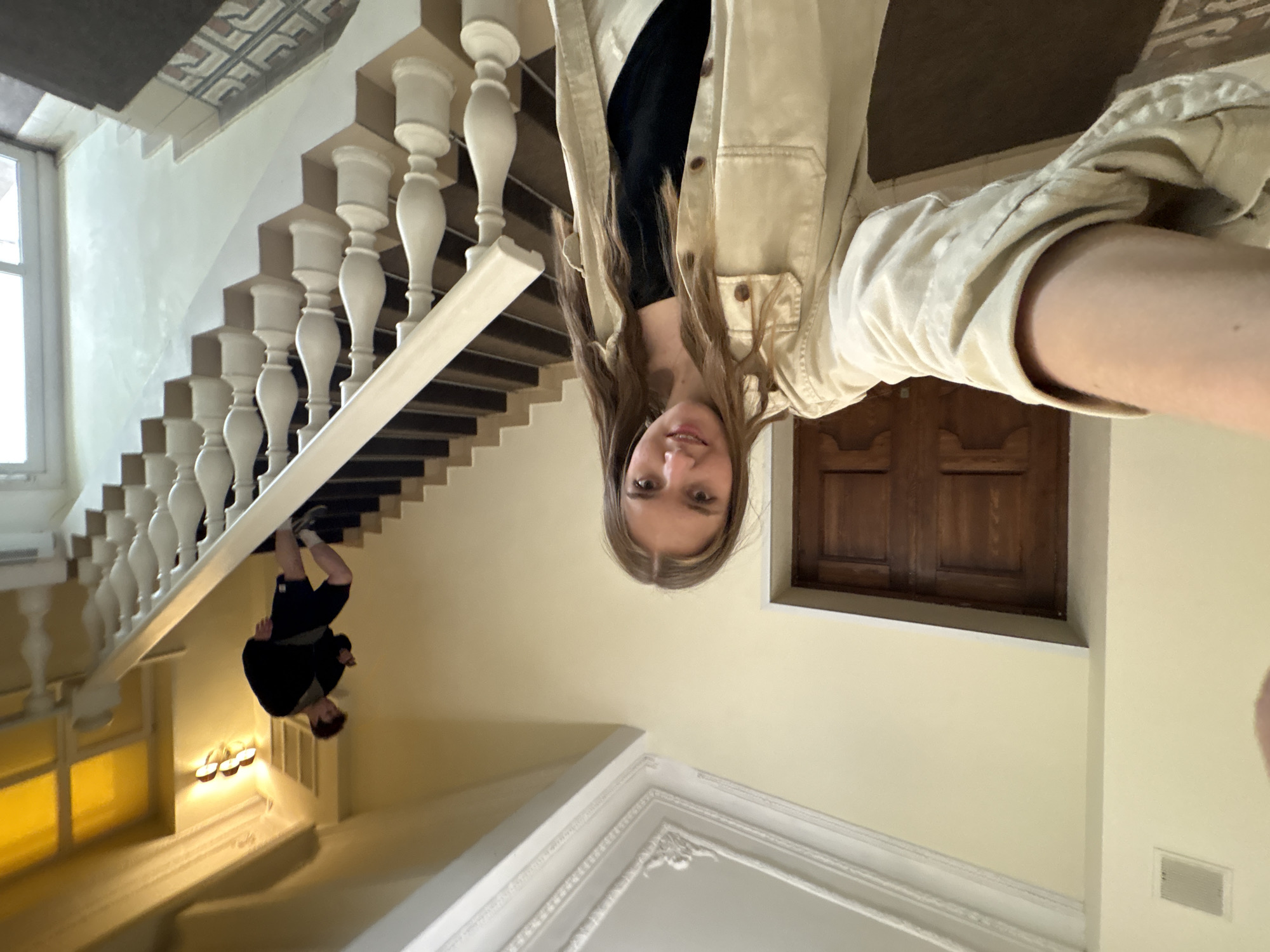 A medium shot depicts a Caucasian woman, positioned on the right, presumably holding a smartphone, taking a selfie. The image is rotated counterclockwise approximately 90 degrees. Behind her is a staircase with white balusters. Another person, a young boy, is upside-down on the staircase. A wooden window is on the right. The location is Perm, Russia.